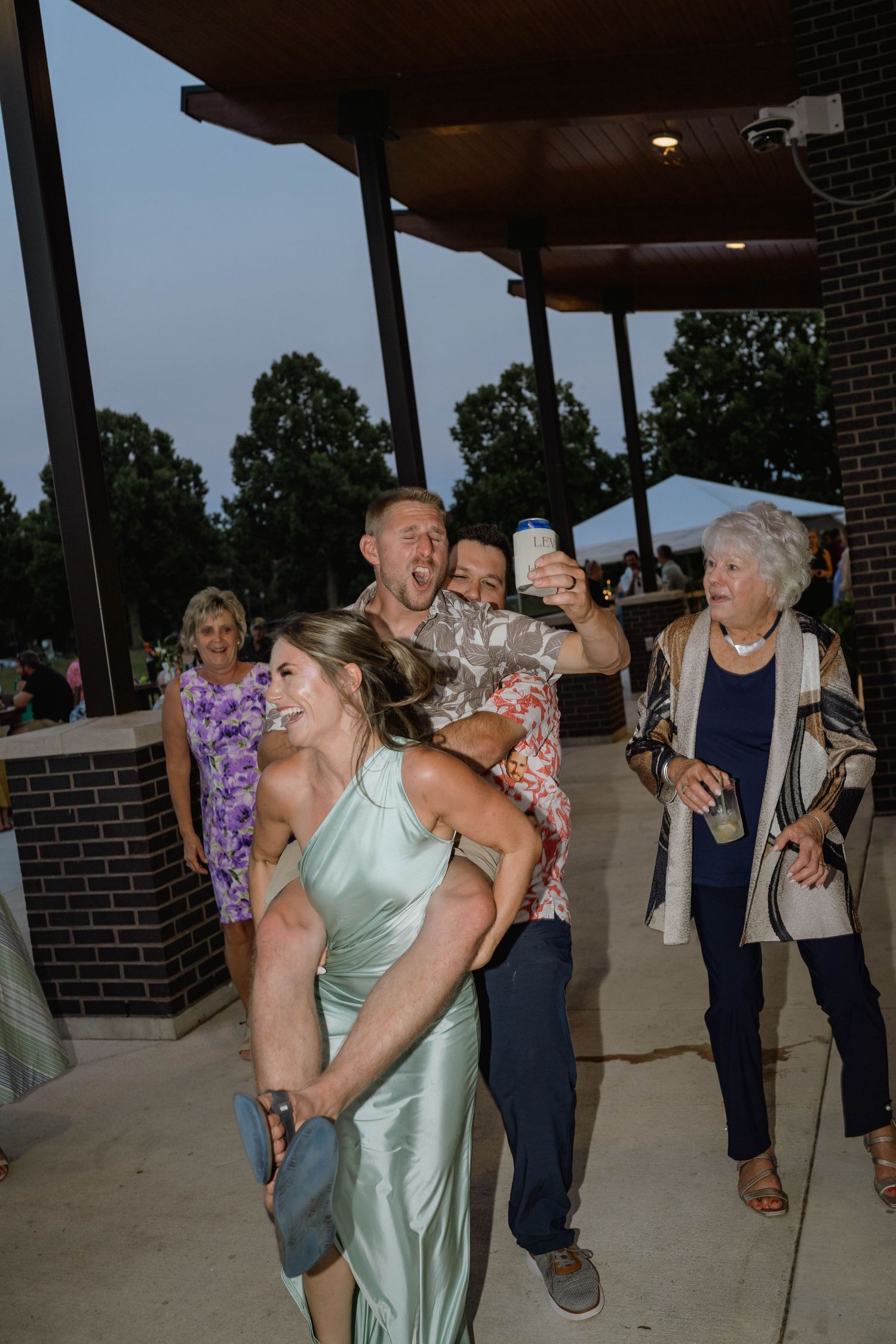 Wedding guests celebrate as man in floral shirt gives bridesmaid a piggyback ride at reception.