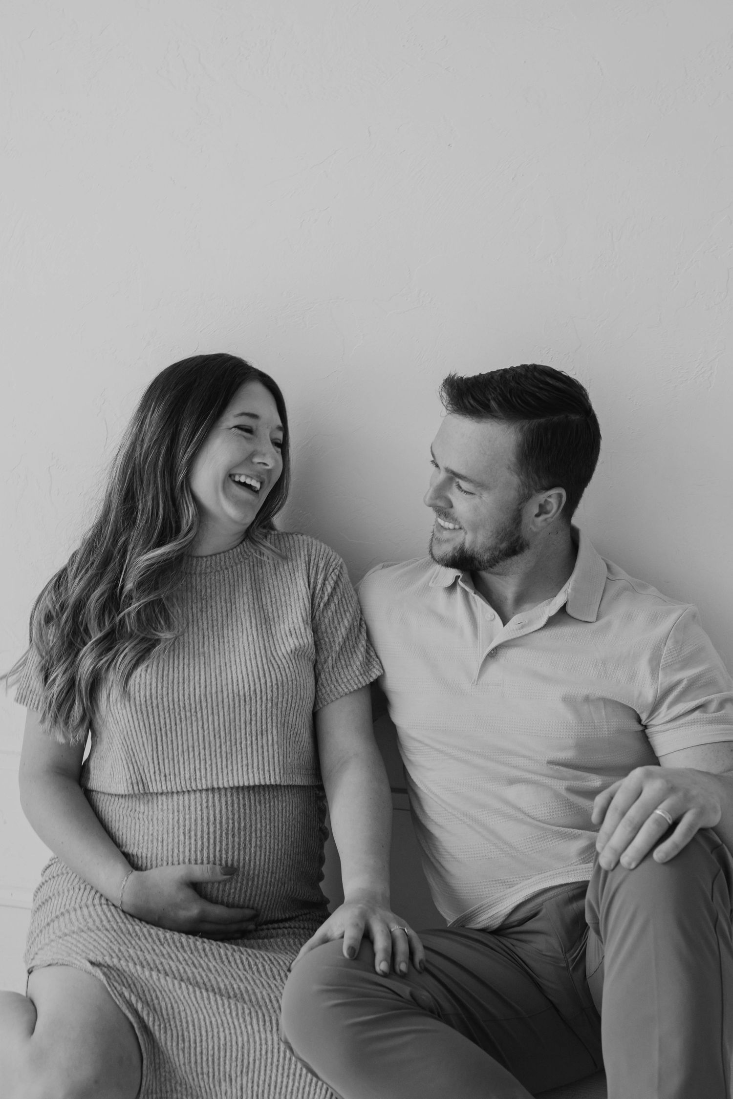 Black and white maternity photo of laughing pregnant woman and smiling partner sitting against white wall.