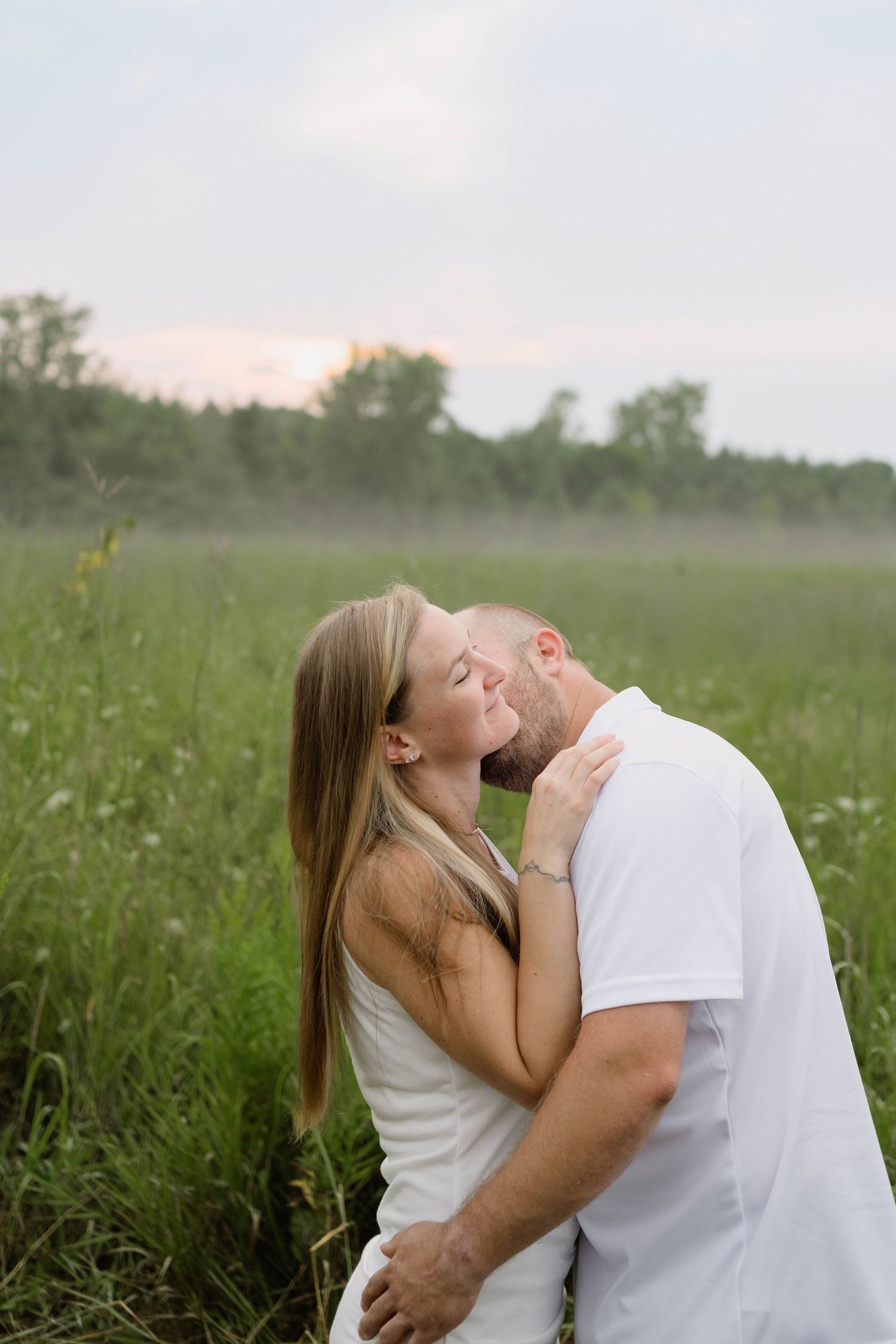 Engaged couple sharing a romantic kiss in a misty green meadow at dusk, both dressed in white.
