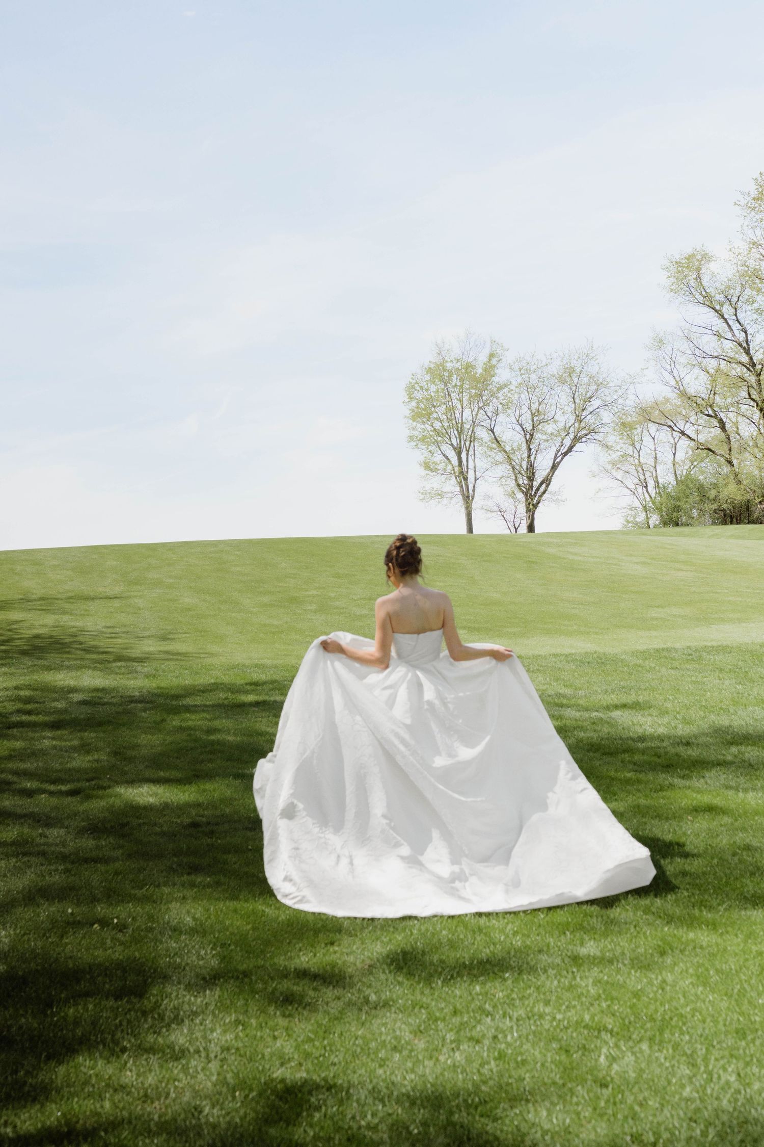 Bride in a flowing white wedding gown walks across lush green grass with trees in the background.
