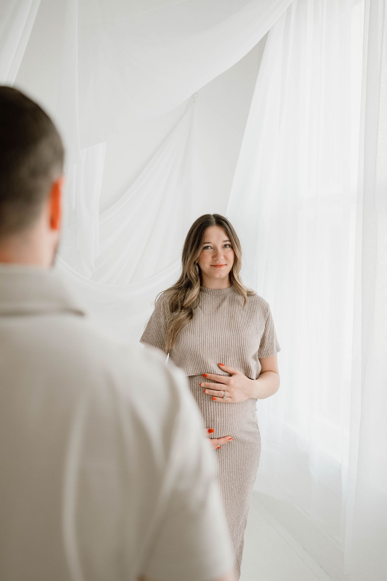 Expectant mother in ribbed beige dress cradling bump, smiling at partner in white studio.