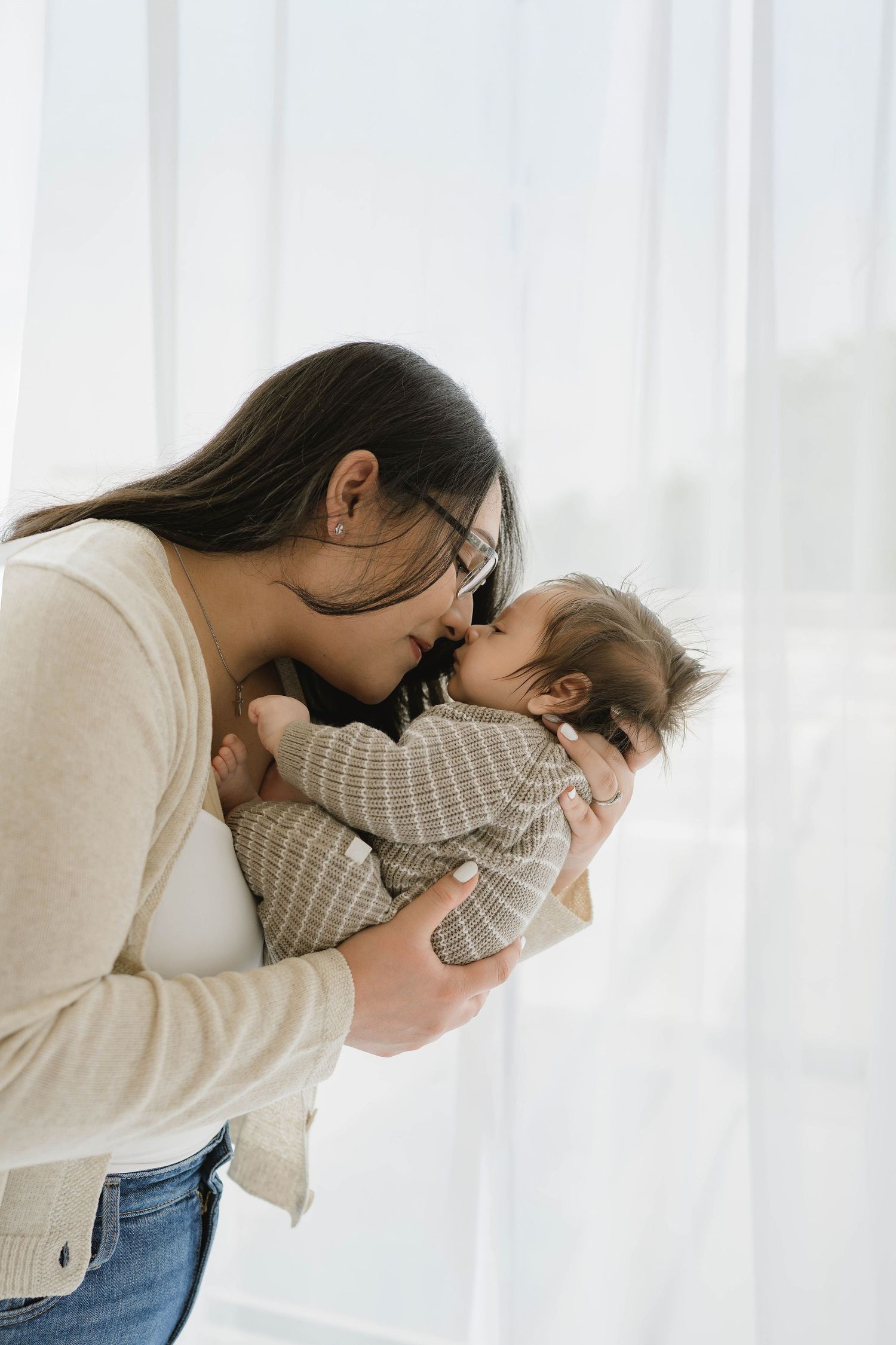 Loving mother wearing glasses tenderly kisses her newborn baby near a bright white curtained window.