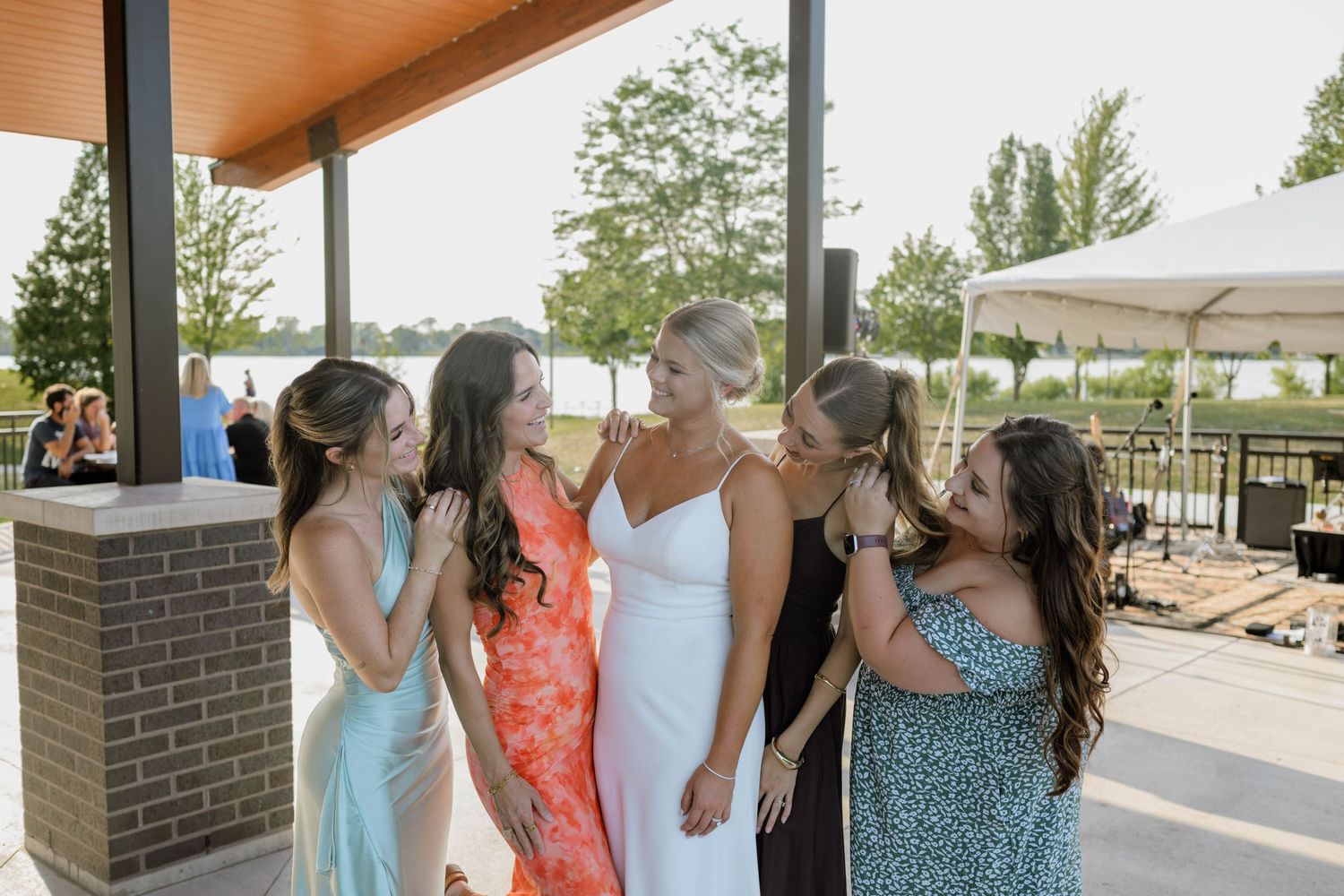 Bride in white dress laughing with bridesmaids in colorful gowns at outdoor wedding reception near waterfront.