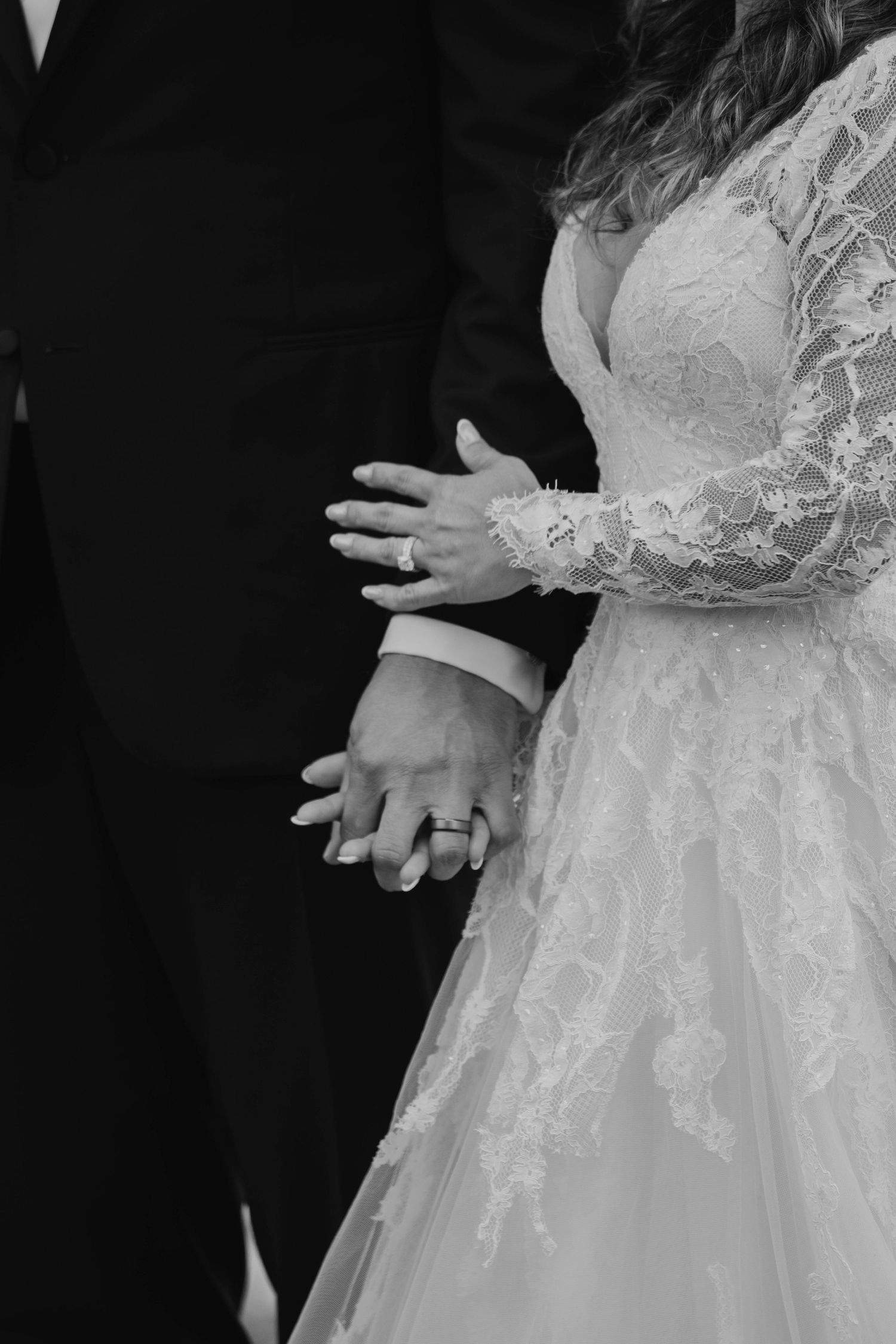 Black and white close-up of bride and groom holding hands showing wedding rings in lace gown.