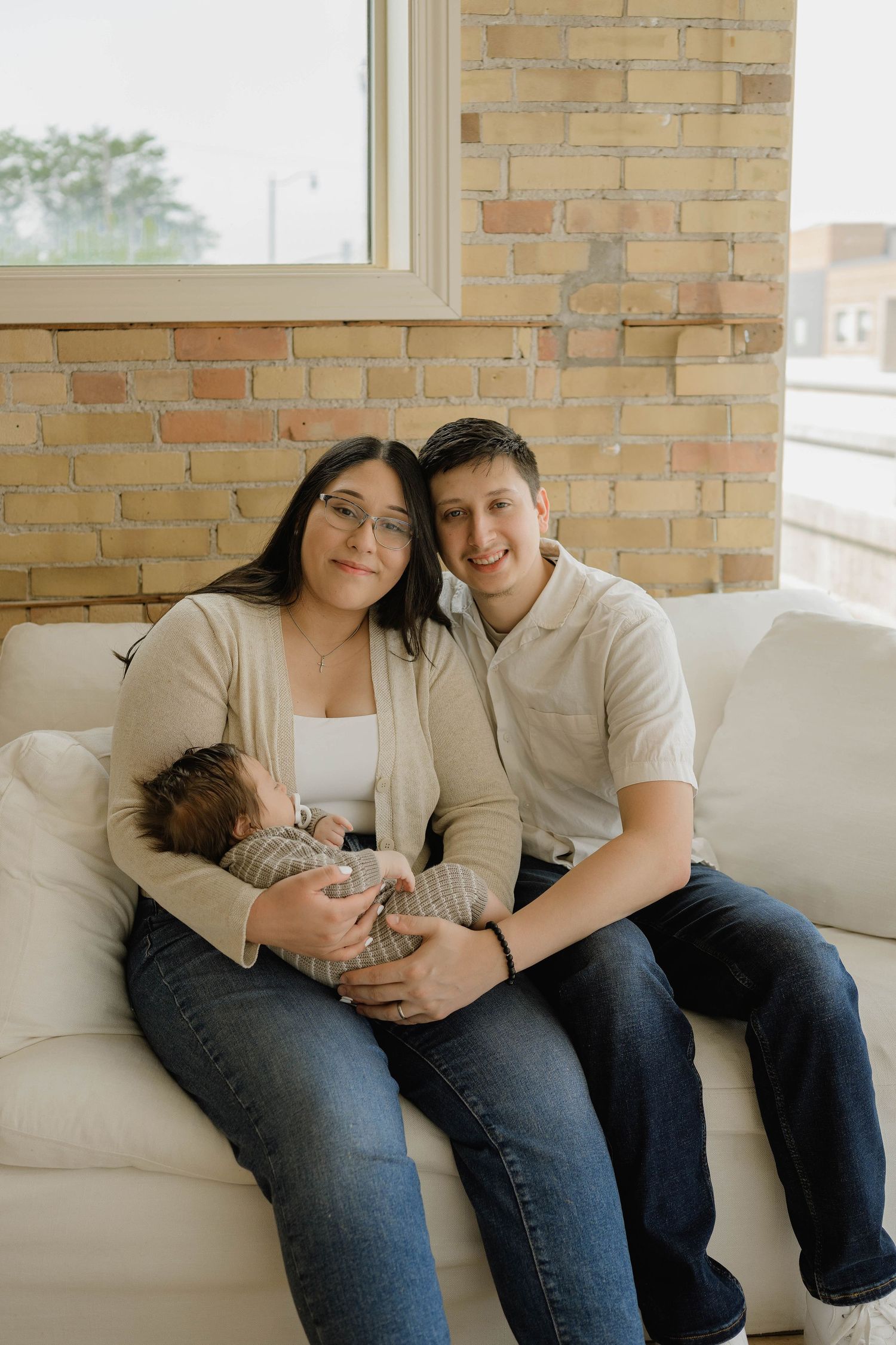 Happy young couple sitting on white sofa holding their newborn baby in a brick-walled home.