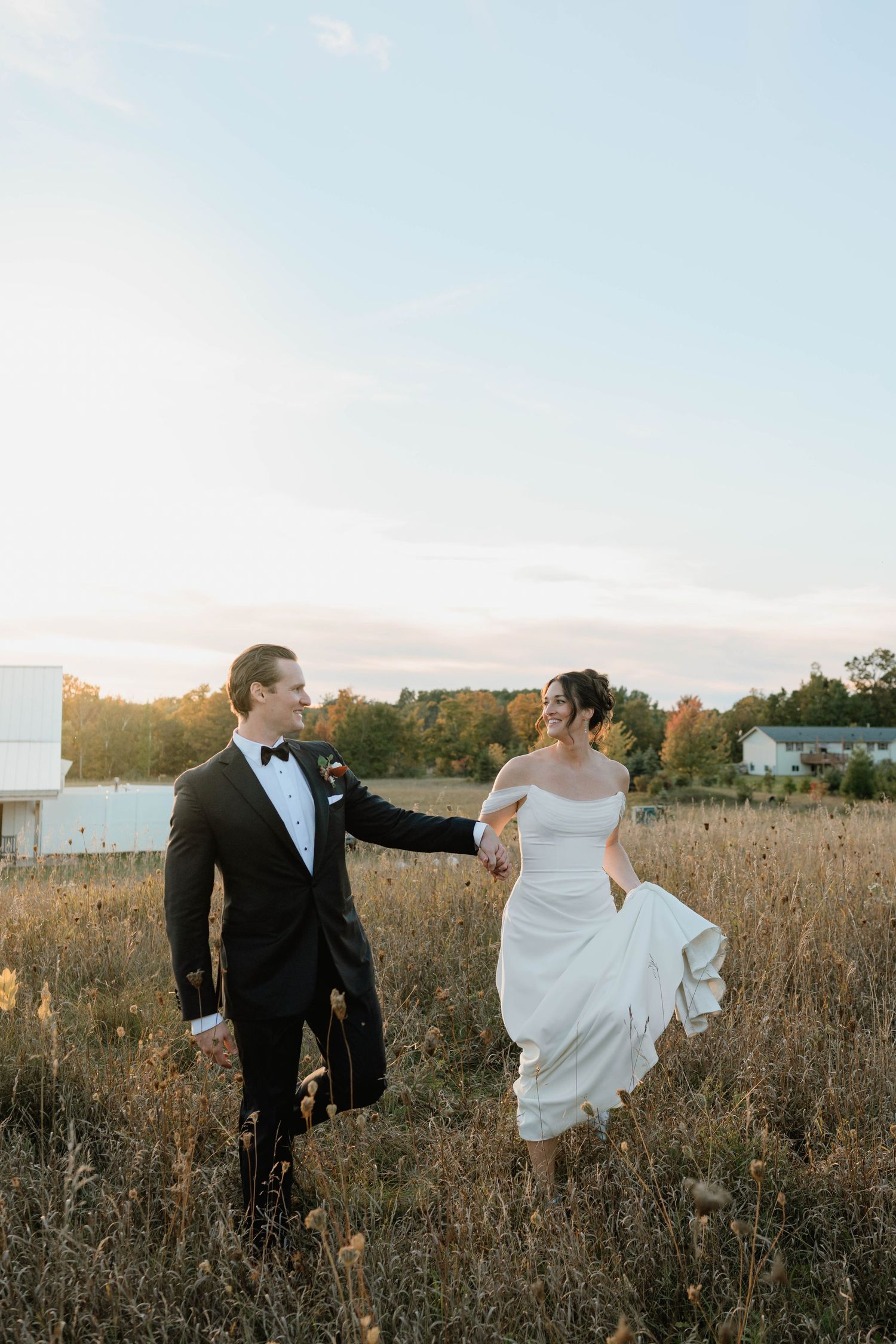 Bride and groom holding hands walking through golden meadow at sunset, she wears off-shoulder gown, he wears black tuxedo.