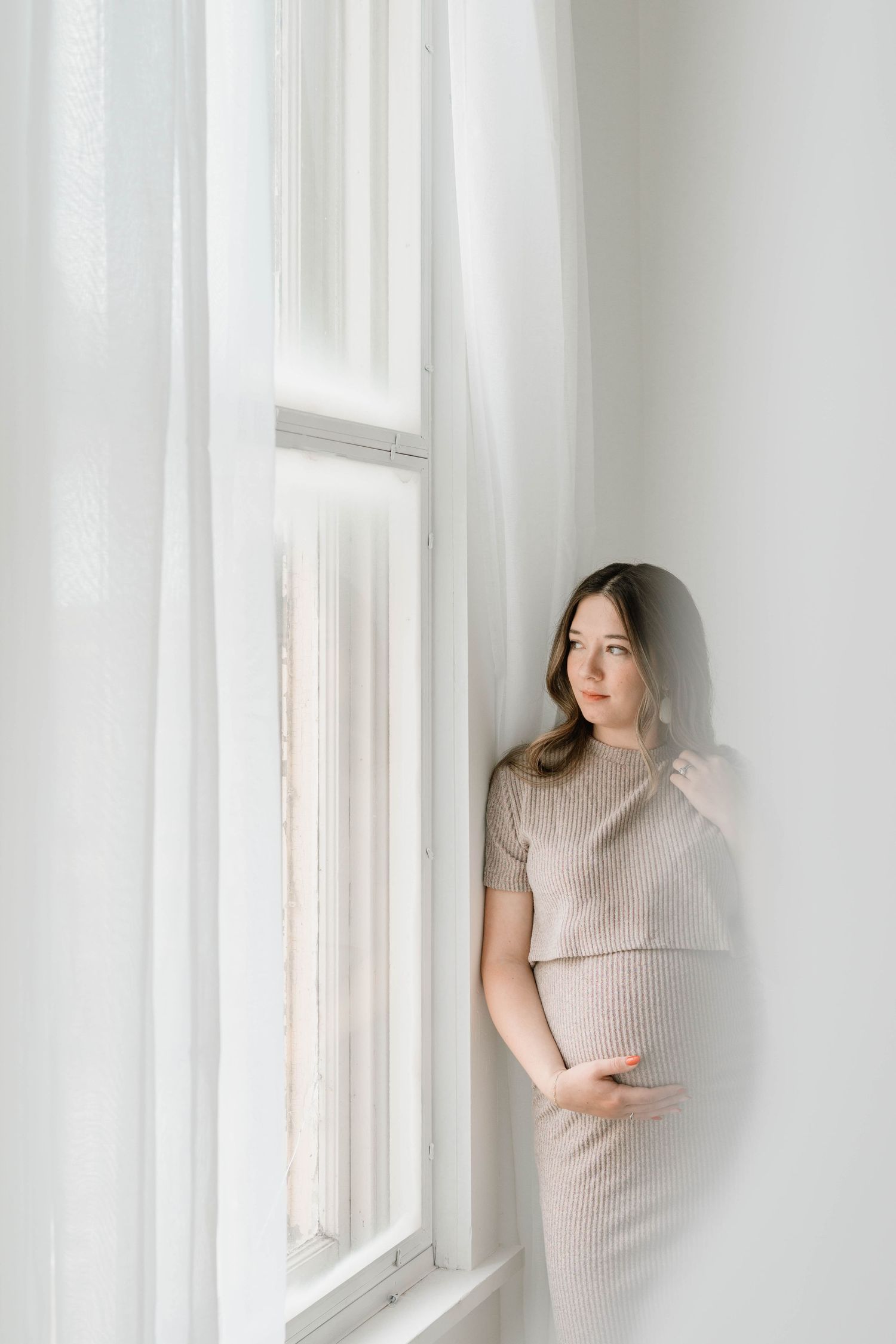 Pregnant woman in beige ribbed dress standing by white window with sheer curtains.