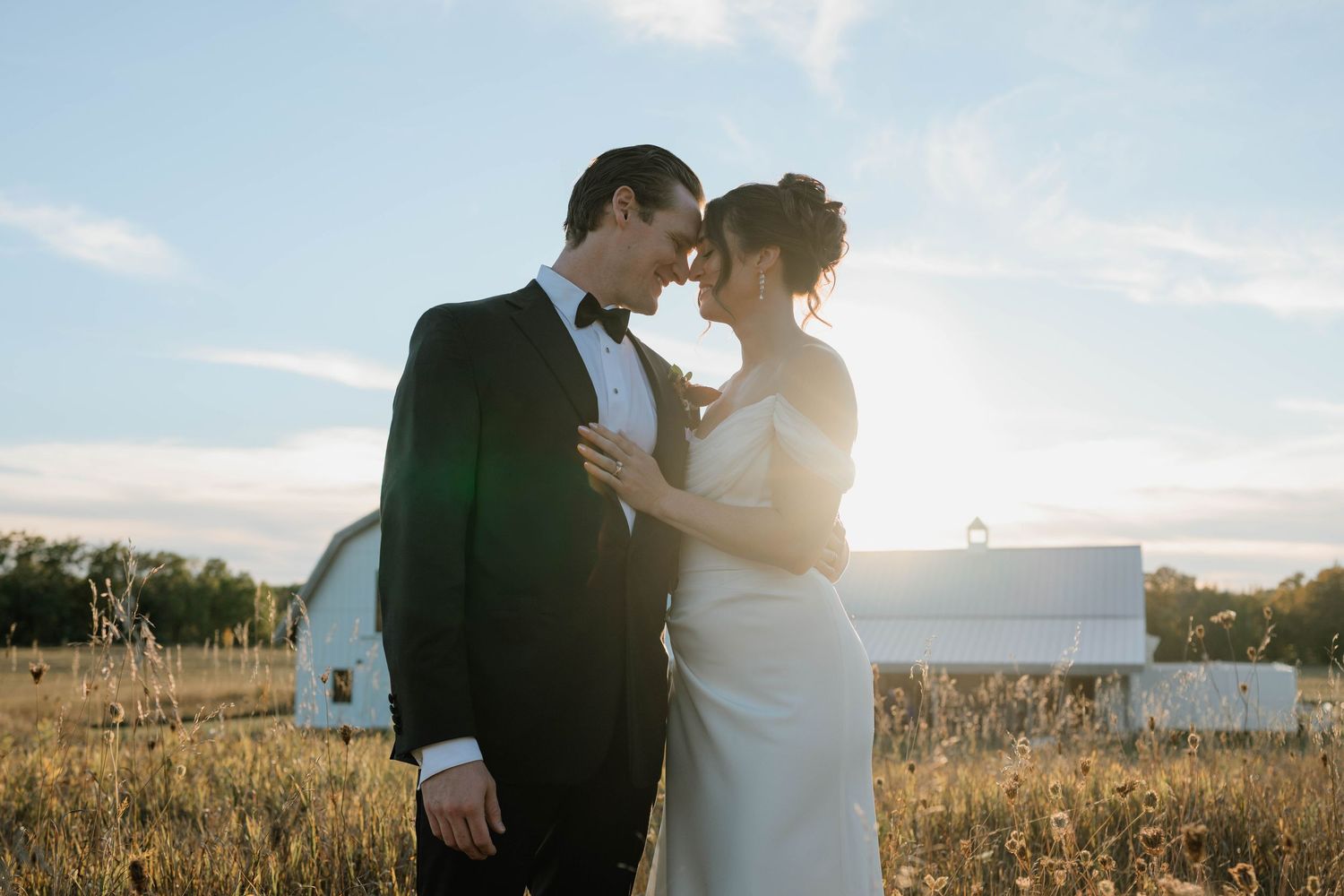 A bride and groom share a romantic moment in a golden field with a white barn at sunset on their wedding day.