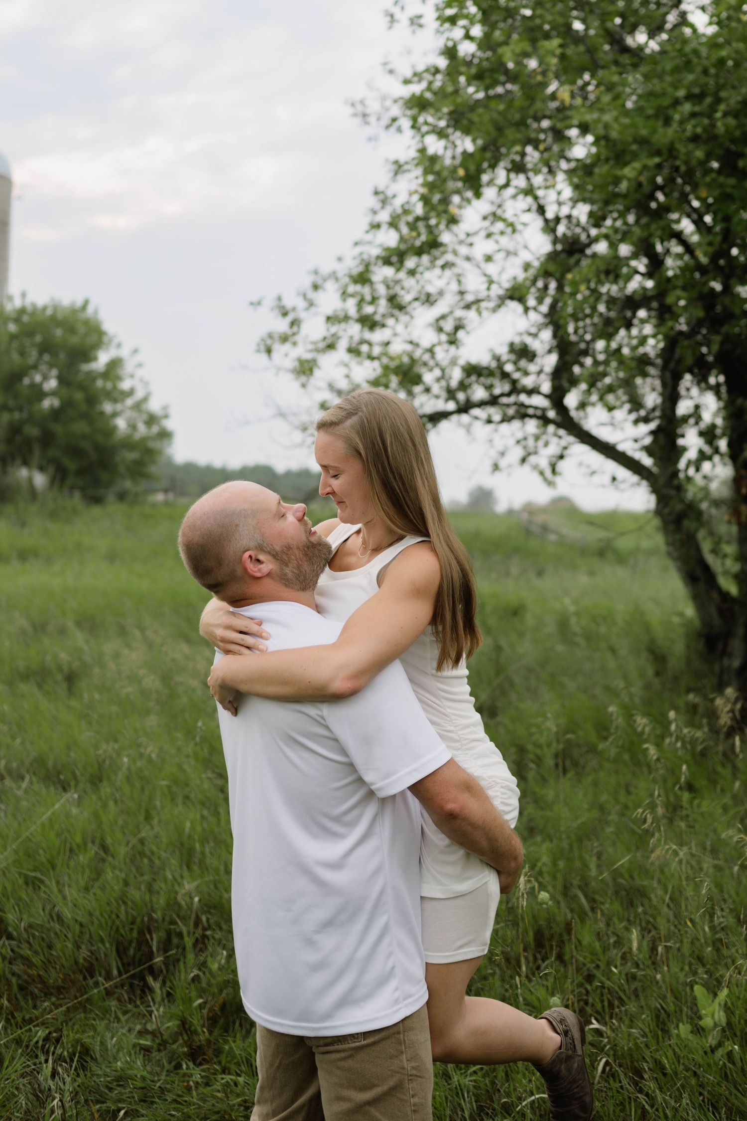 Man lifts woman in white dress while laughing together in green meadow during engagement session.