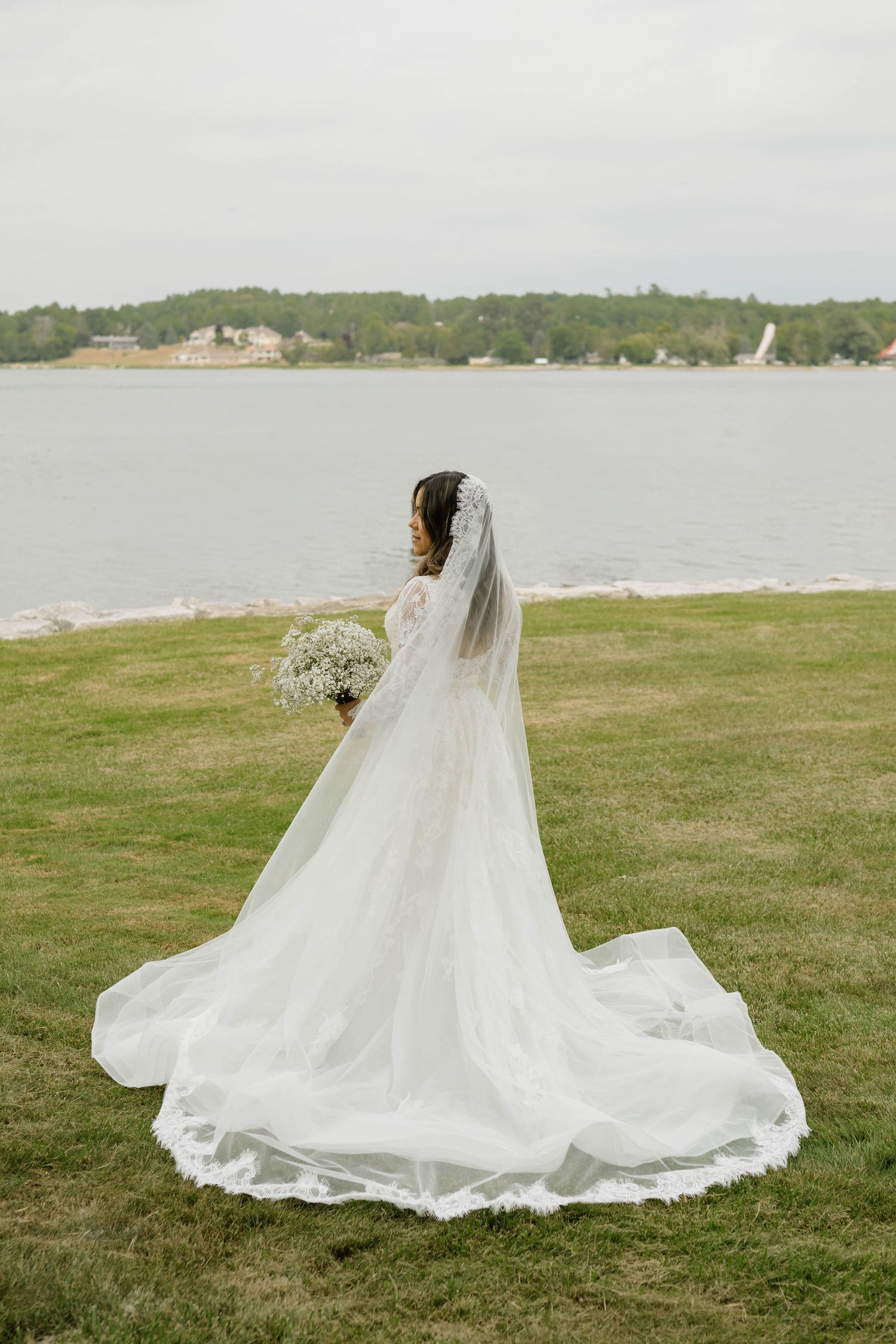 Bride in a long white wedding gown with veil and bouquet standing on green grass by a serene waterfront.