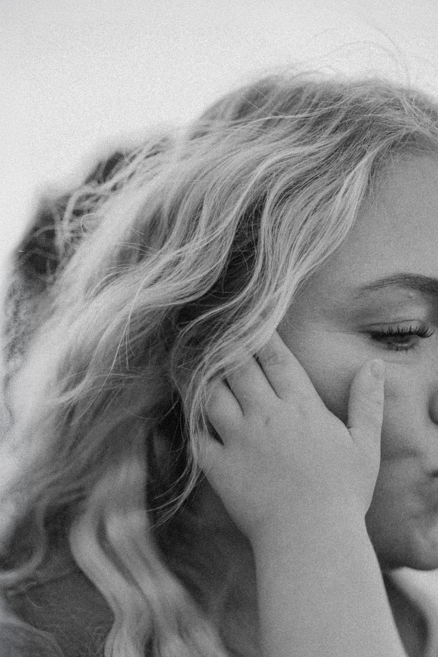 Black and white portrait of a blonde woman with wavy hair, eyes closed, hand raised to her face.