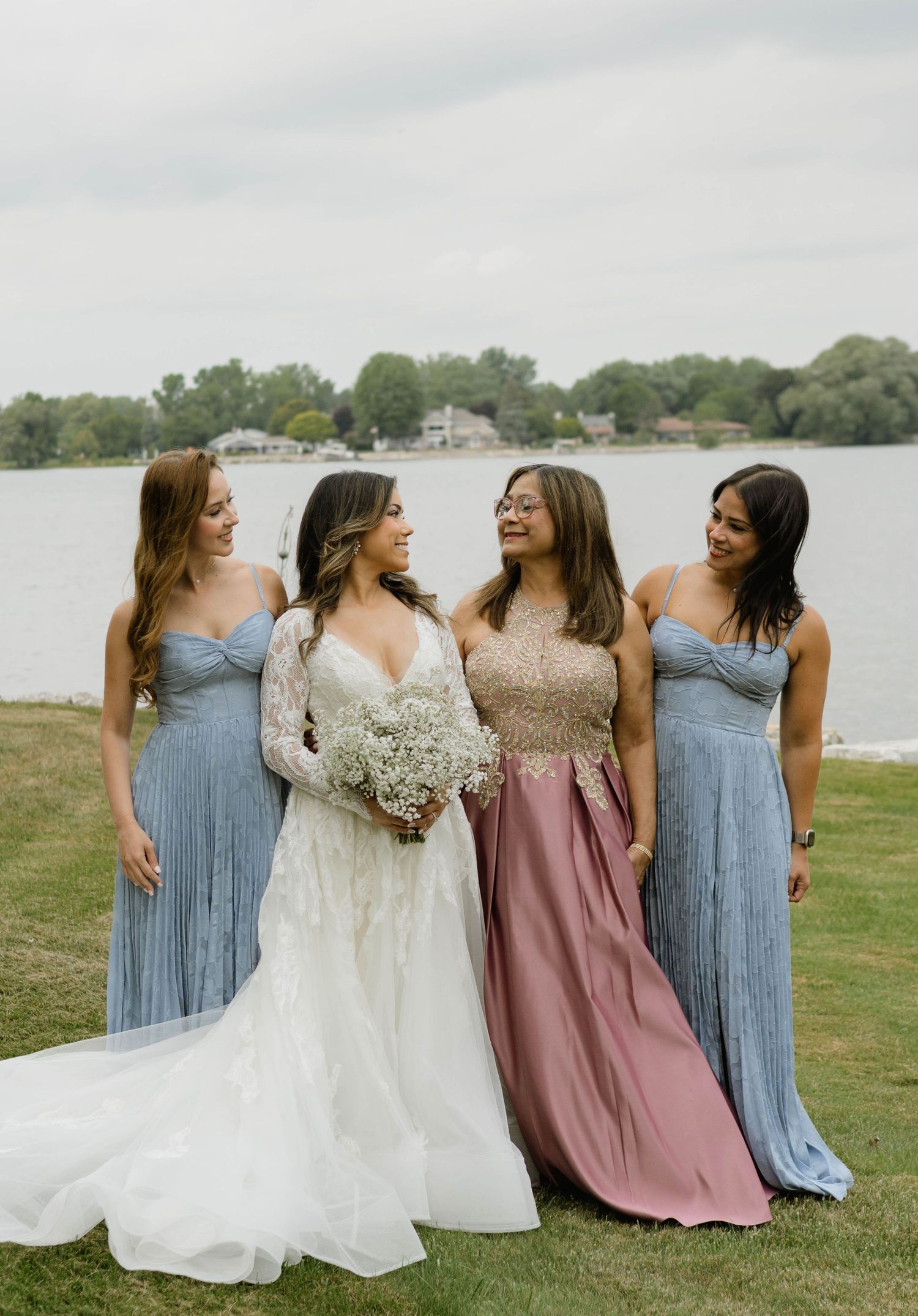 Bride in lace gown holding white bouquet with three bridesmaids in blue and mauve dresses by lake.