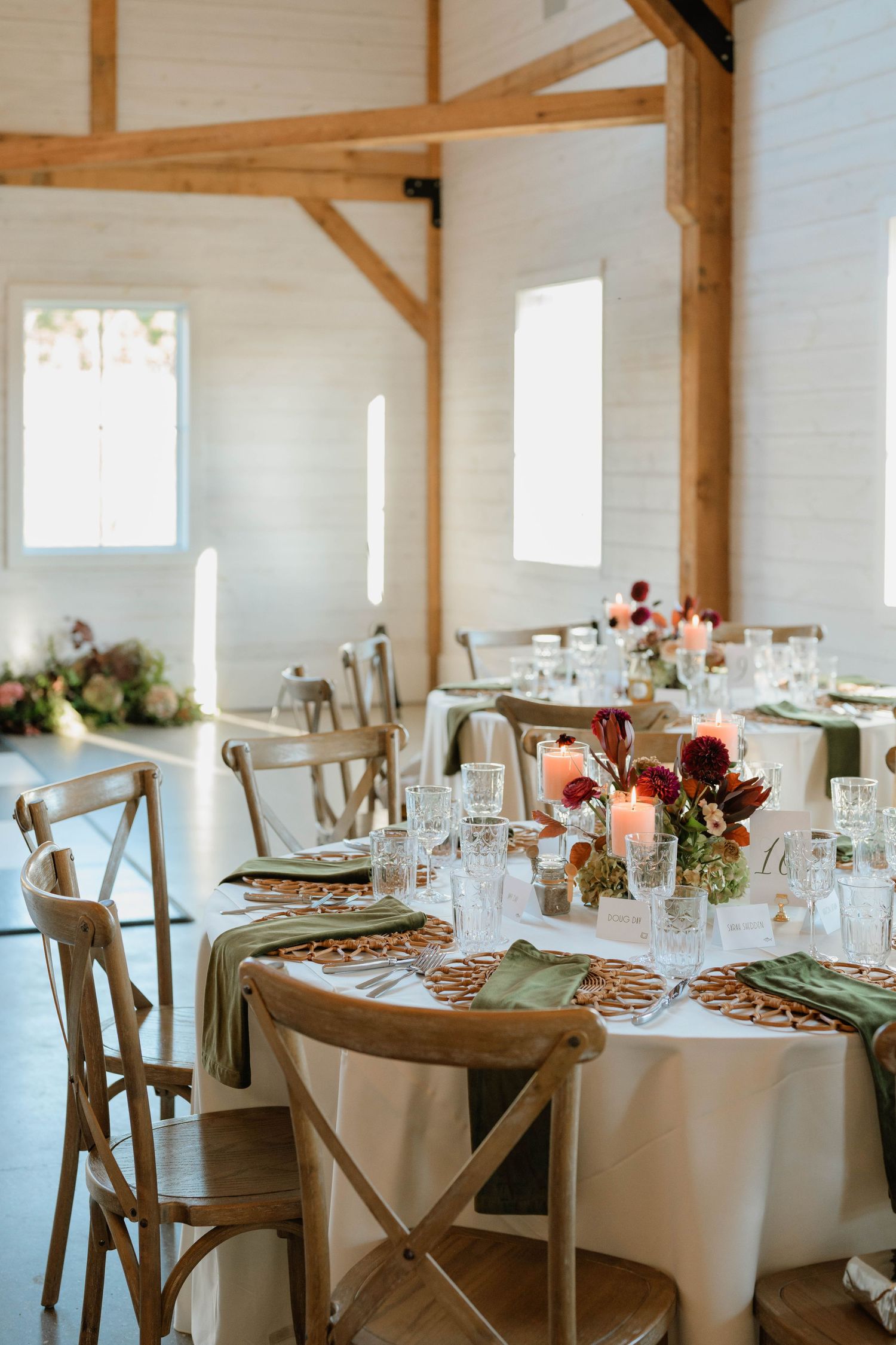 Elegant barn wedding reception with wooden cross-back chairs, green napkins, floral centerpieces, and candles on white linen tables.