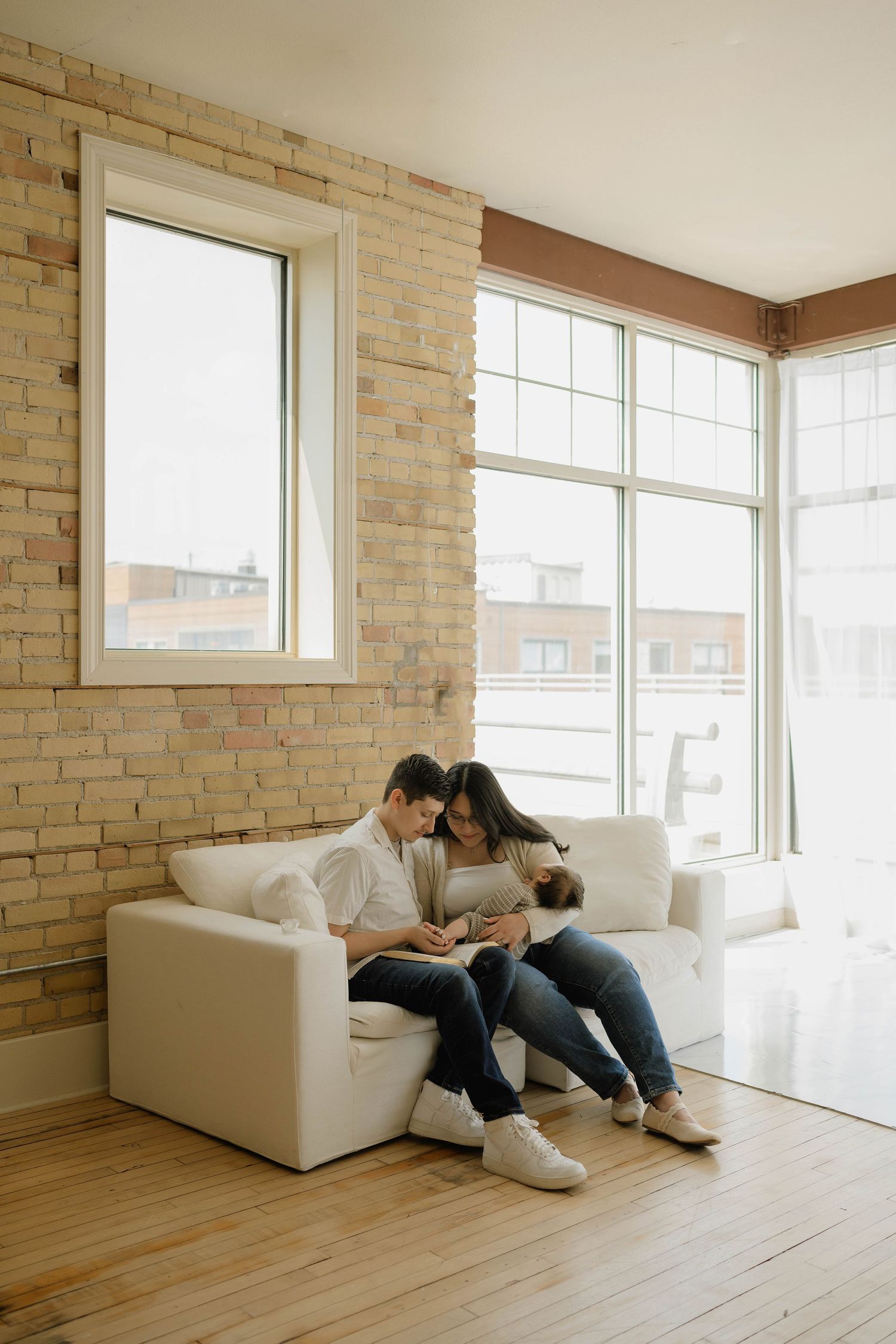 Young couple sitting together on a white couch admiring their newborn baby in a bright loft home.