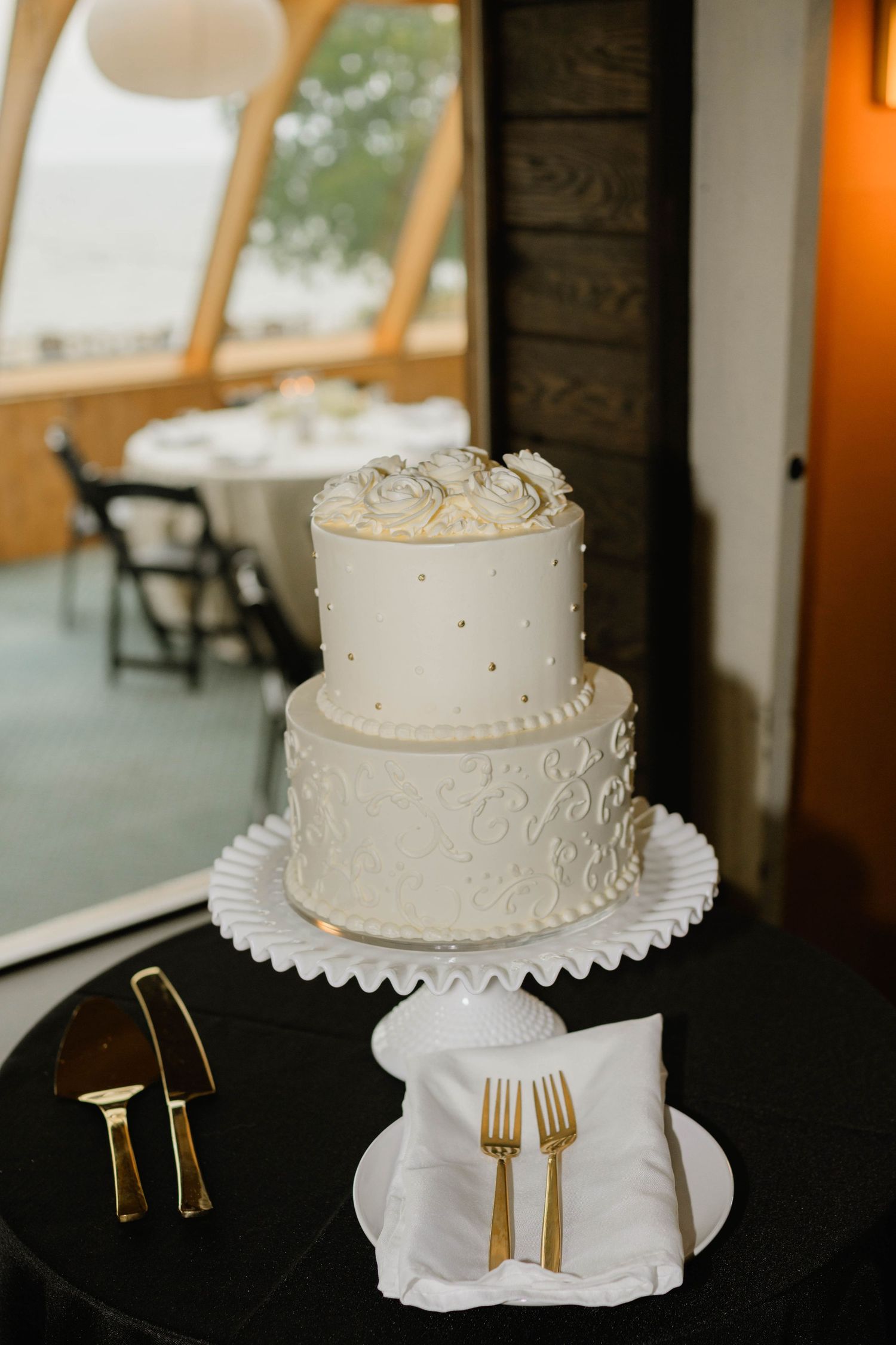 Two-tier white wedding cake with gold dot accents and floral topper on black cake stand.