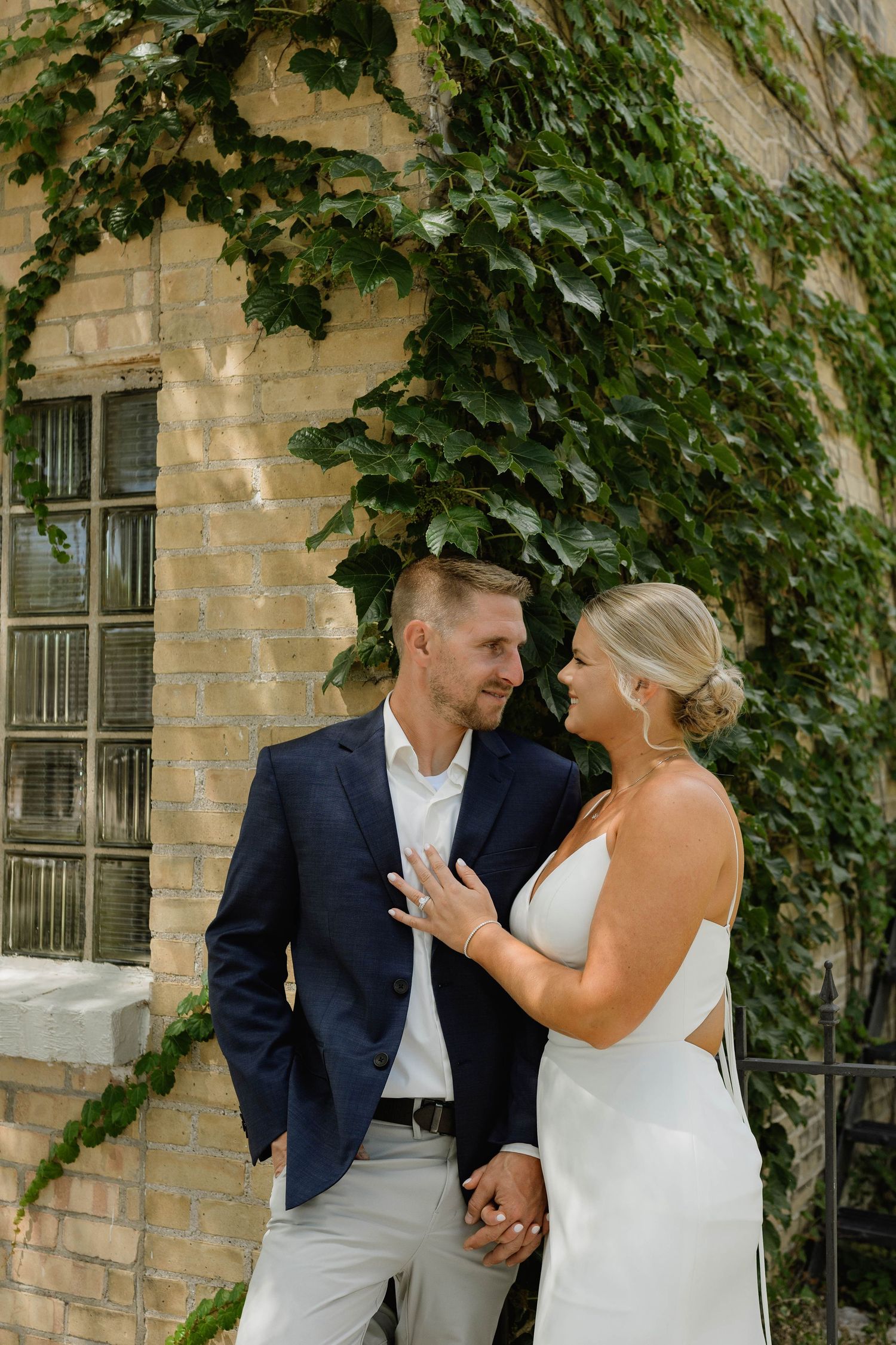Bride and groom smiling at each other while holding hands in front of ivy-covered brick wall.