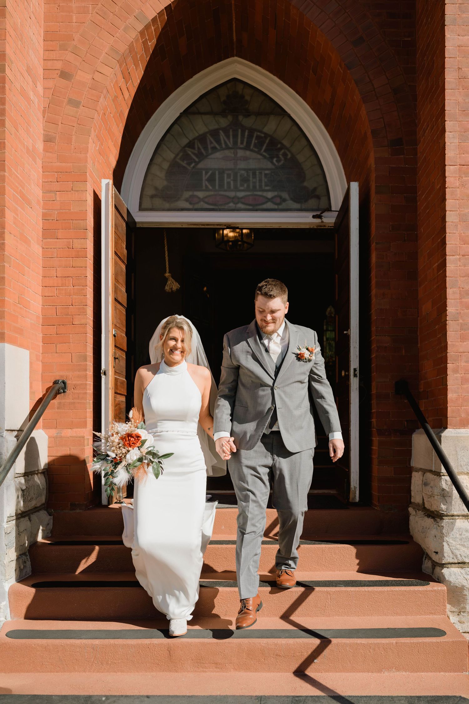 Bride in a white gown with bouquet and groom in a gray suit hold hands as they exit a red brick church after their wedding ceremony.