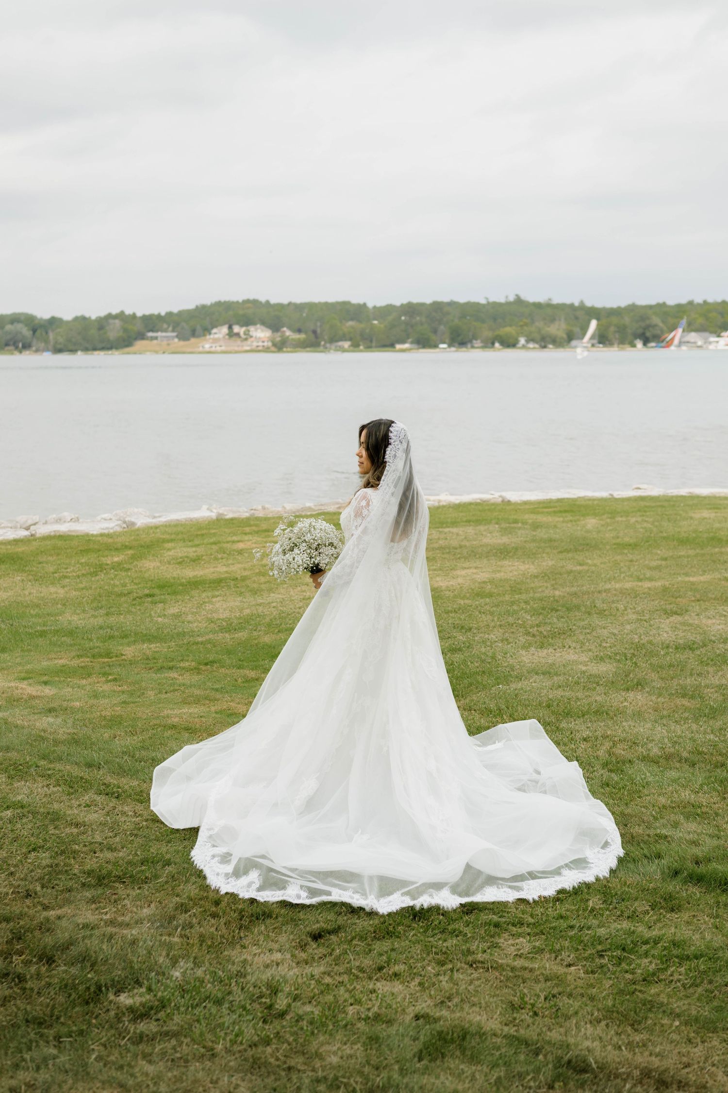 Bride in white gown with cathedral veil holding bouquet standing by a lake on green grass.