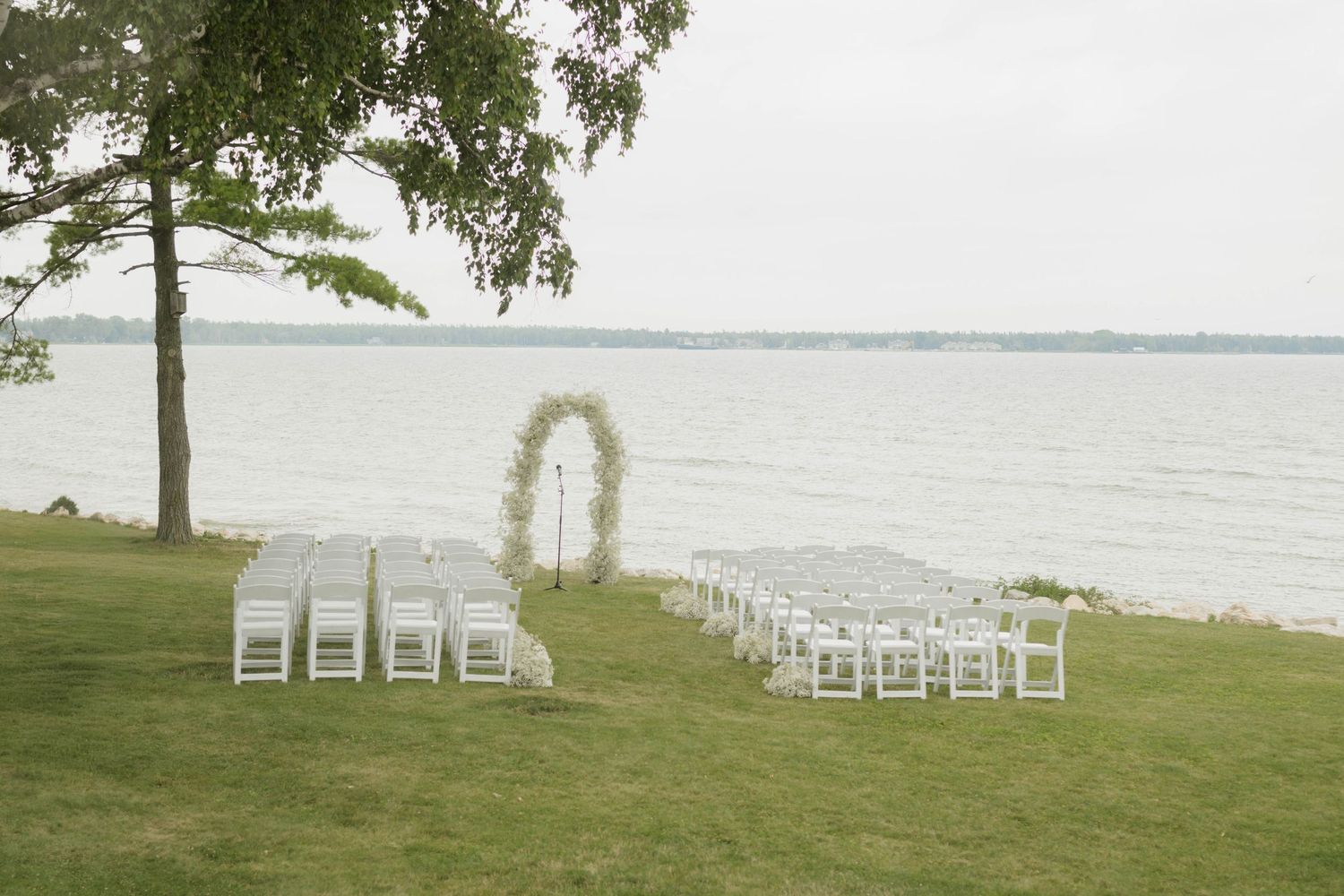 Outdoor lakeside wedding ceremony setup with white chairs and floral arch by the water.