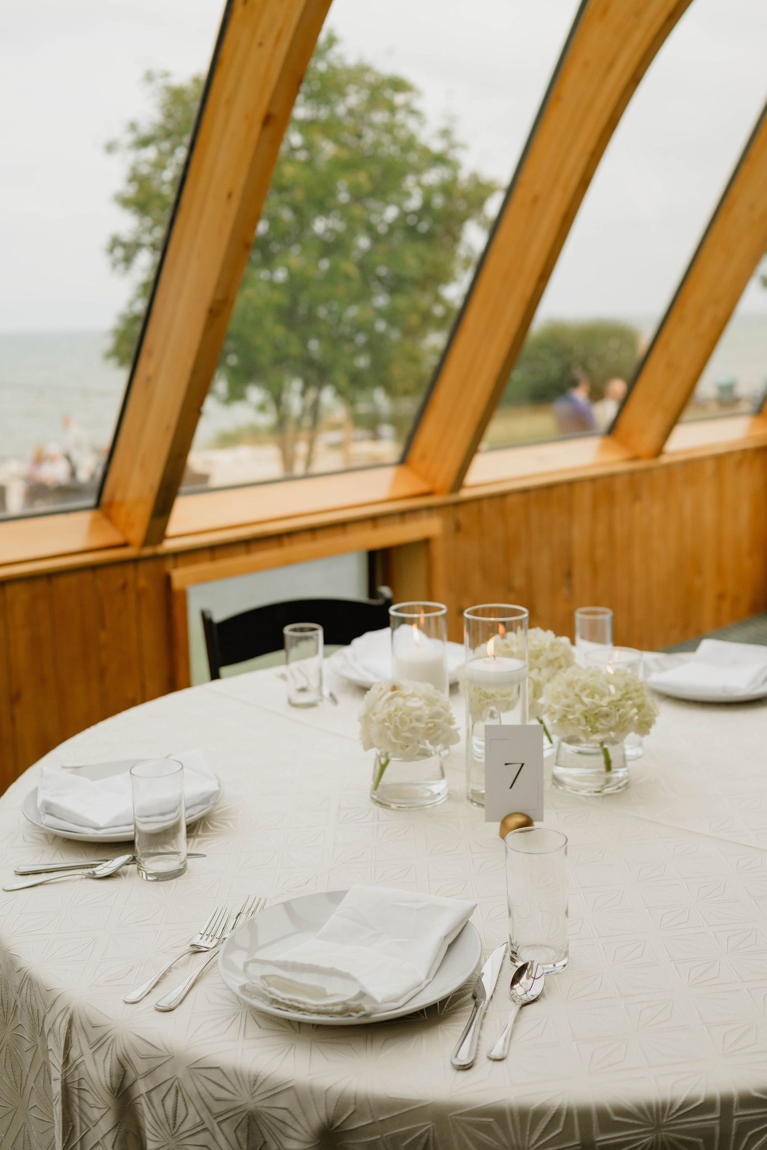 Wedding reception table setting with white florals and table number near large windows overlooking water.