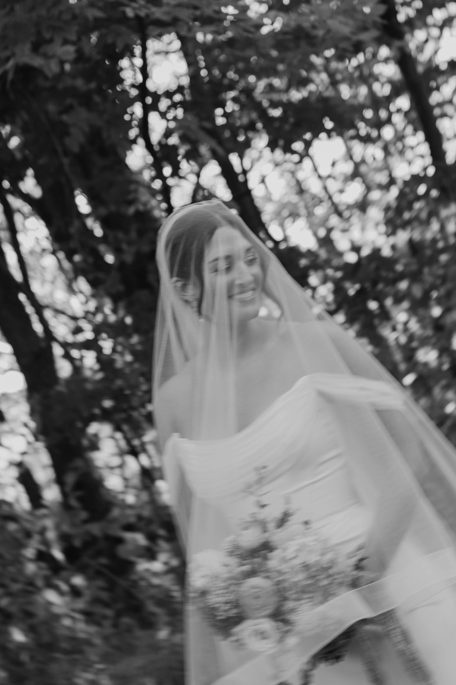 Smiling bride in a flowing veil and strapless gown holds a bouquet among trees in a soft-focus black and white wedding portrait.