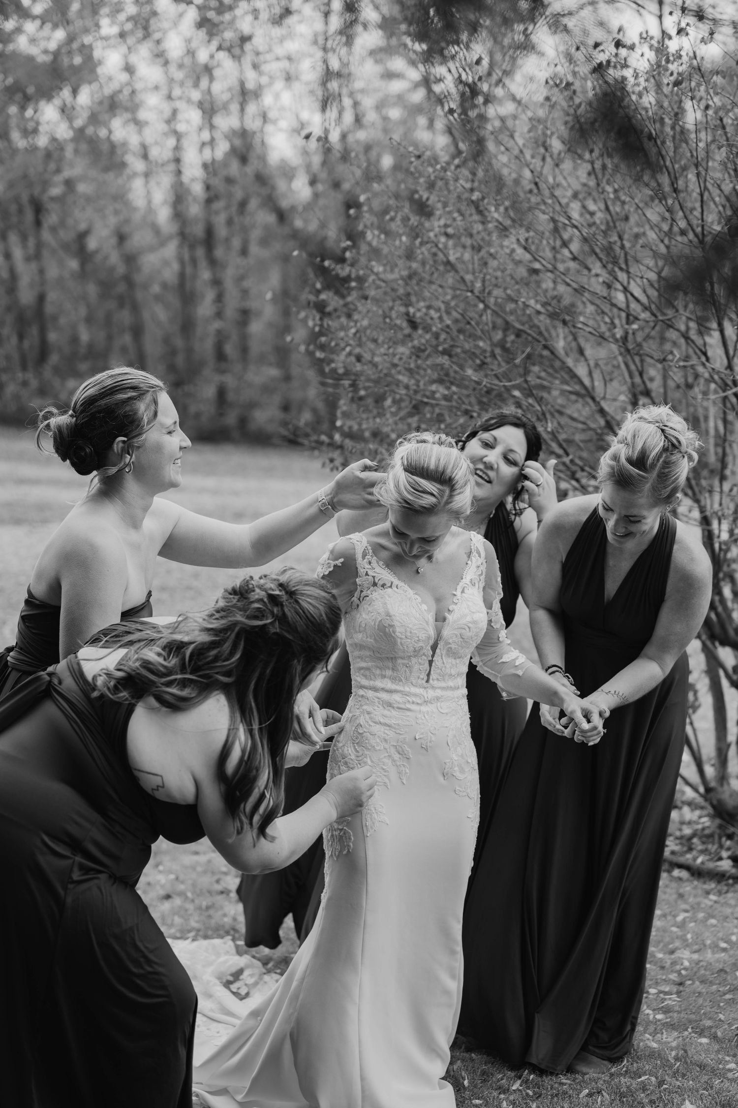 Black and white photo of bridesmaids in dark dresses helping a bride in a lace wedding gown adjust her hair and dress outdoors.