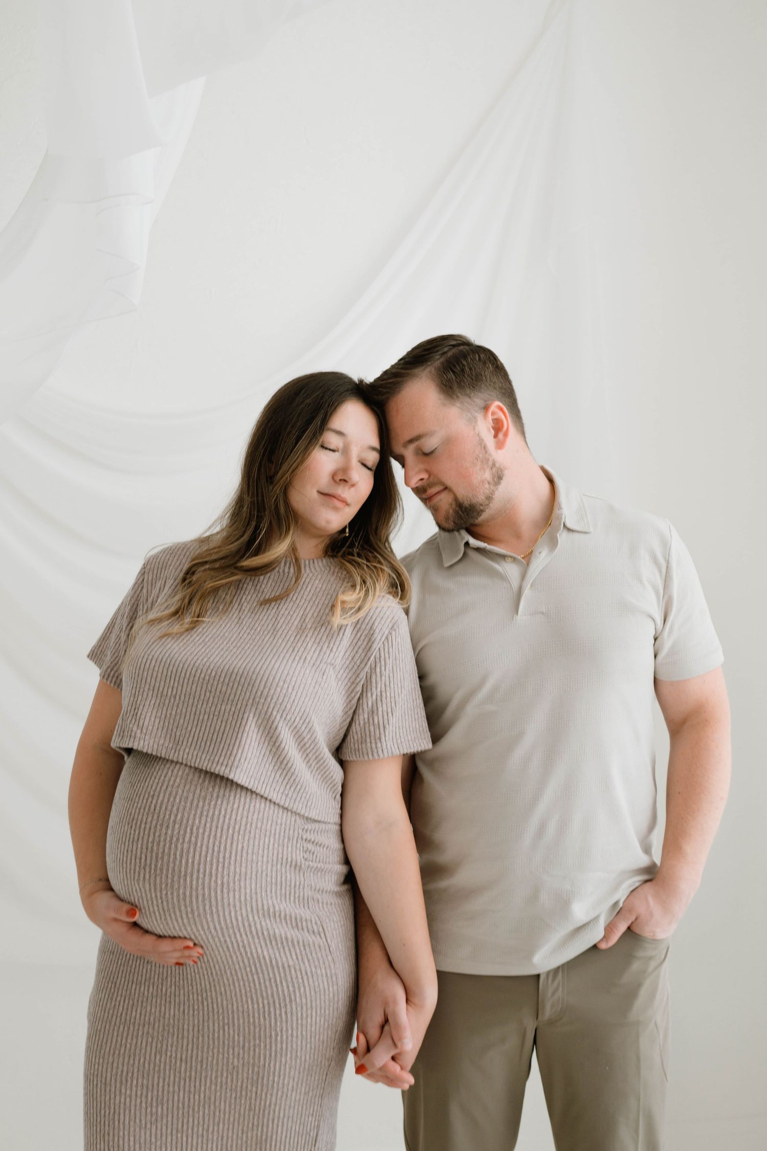 Expecting couple in neutral tones hold hands and lean heads together against white draped backdrop.