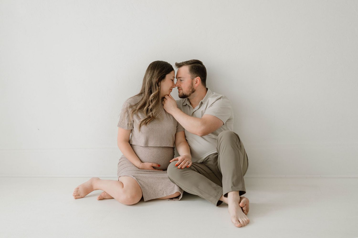 Couple sharing a tender kiss during a maternity photo session on a white studio floor.