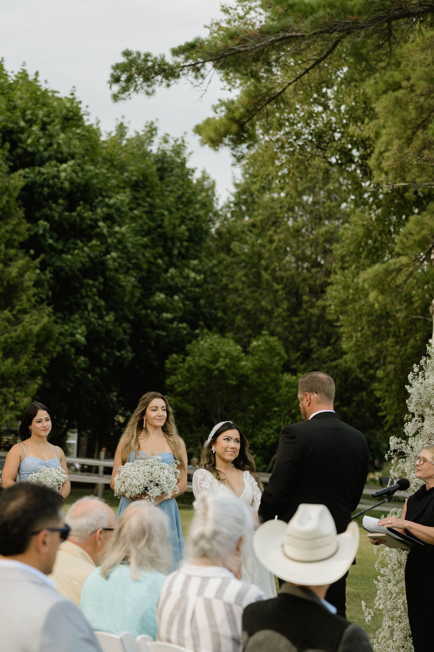 Bride stands with bridesmaids in blue dresses holding baby's breath bouquets at outdoor ceremony.