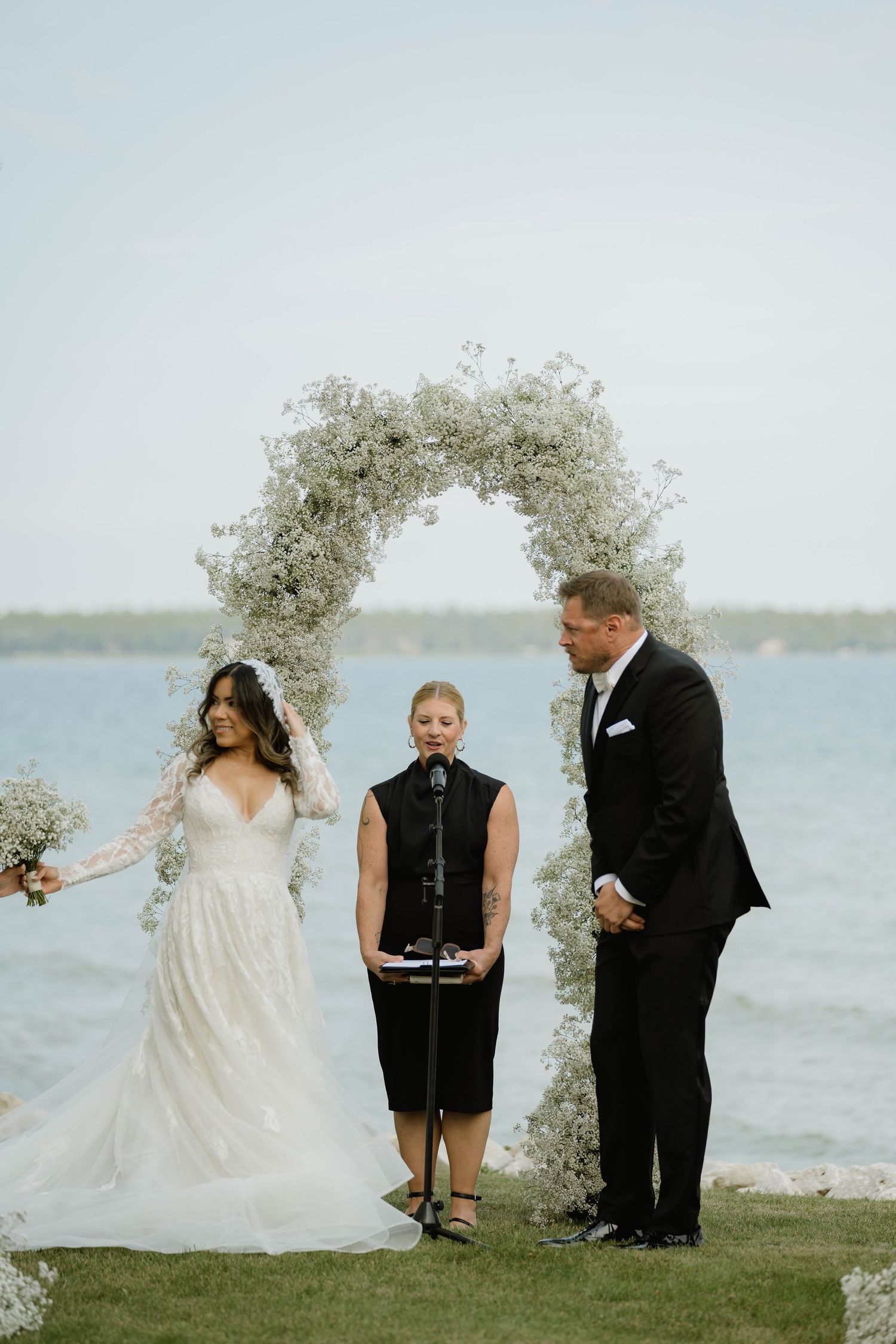Bride and groom exchanging vows under a baby's breath floral arch beside a serene lake.