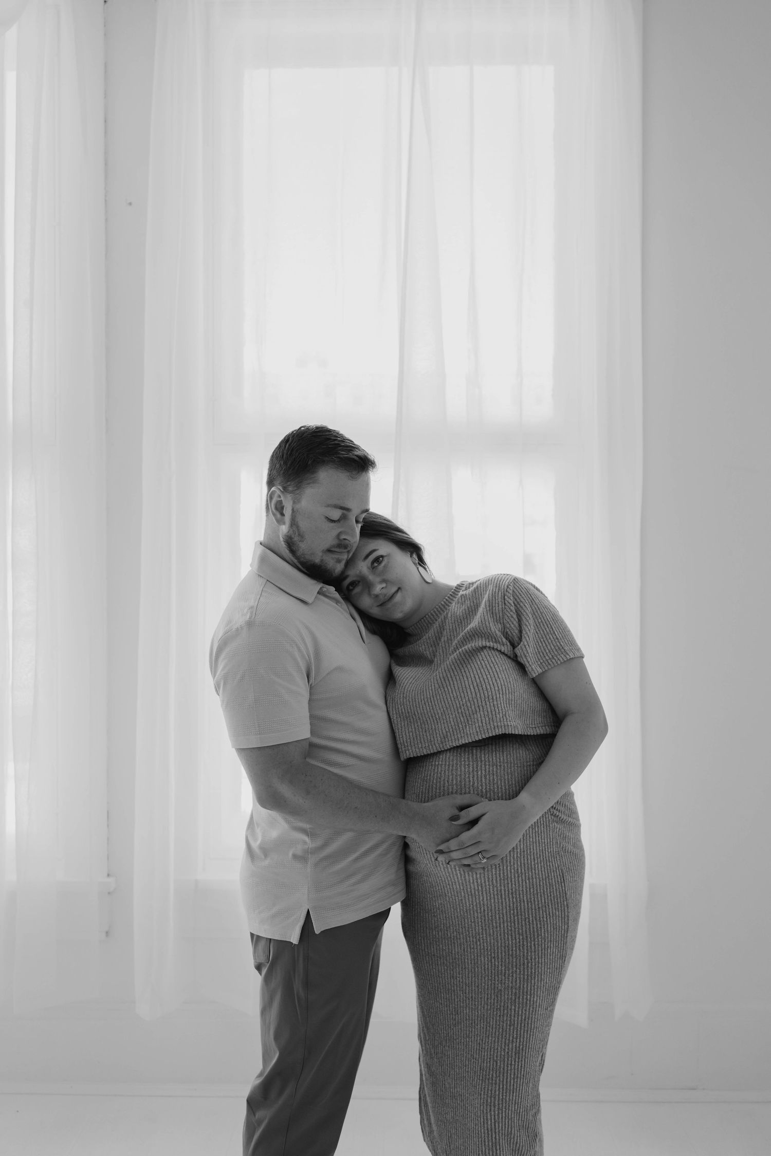 Black and white photo of man tenderly embracing pregnant woman from behind near bright window.