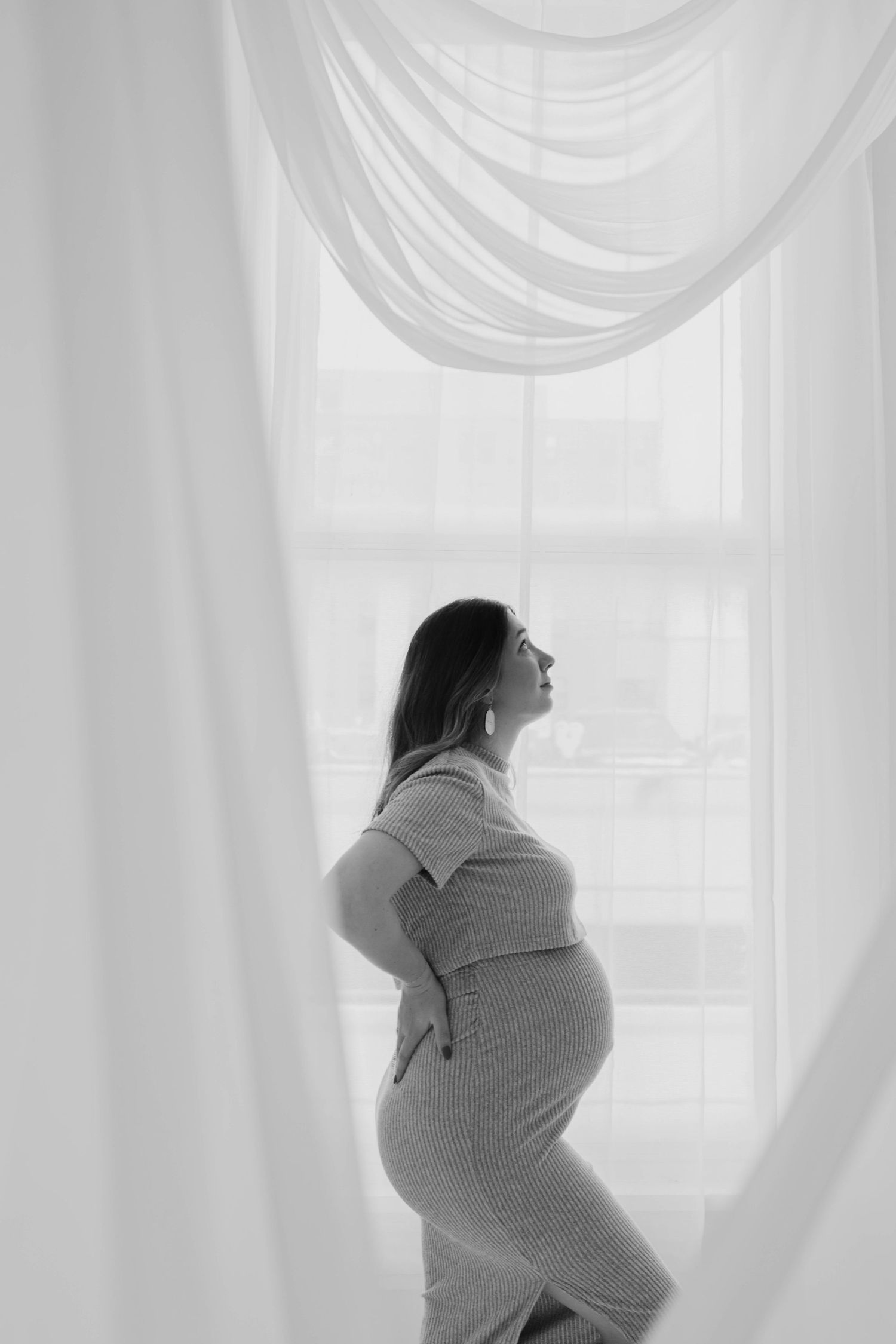Black and white photo of pregnant woman in ribbed dress standing by window with flowing white curtains.