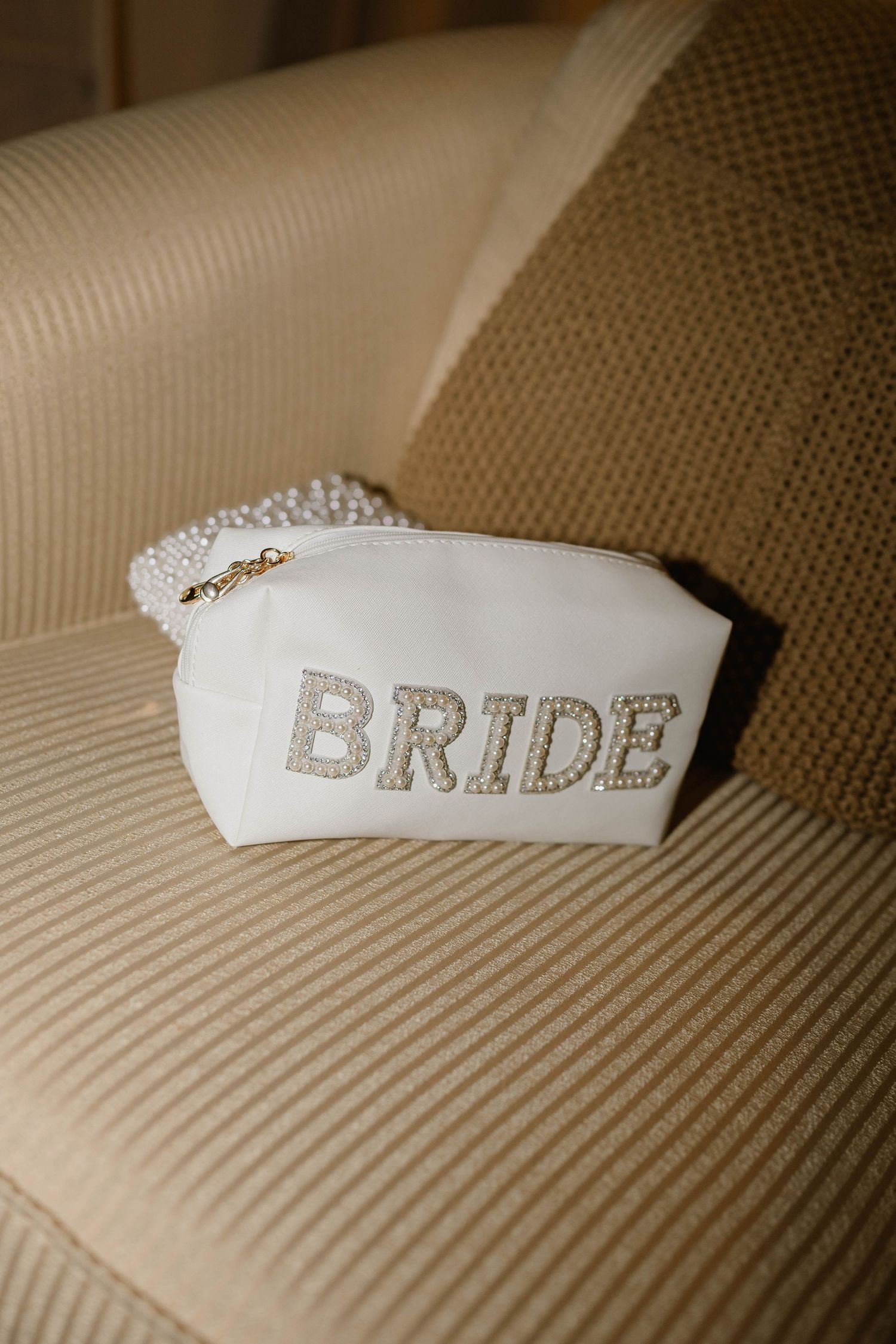 A white pearl-embellished BRIDE cosmetic pouch resting on a beige textured chair.
