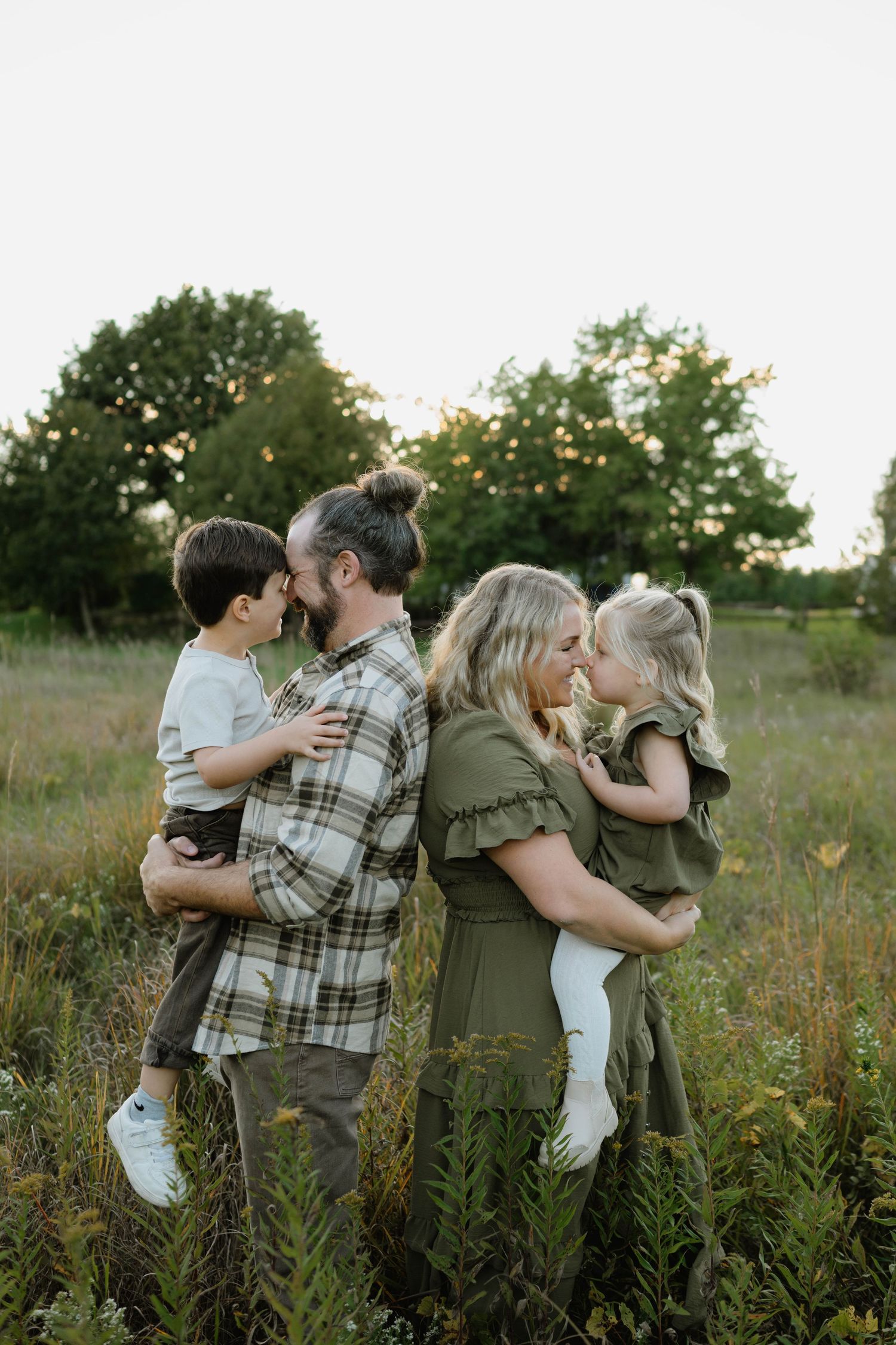 Family of four sharing tender moments in a green field at golden hour, parents holding young children face-to-face.