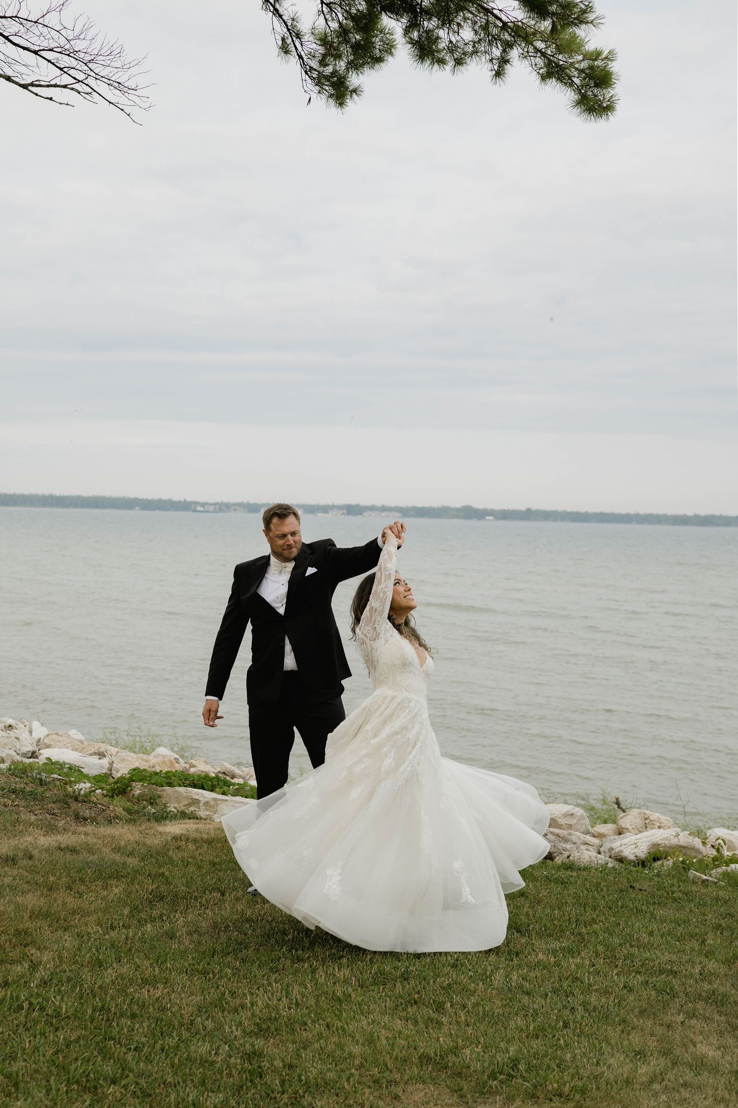 Groom twirling bride in white lace gown by the waterfront on their wedding day.