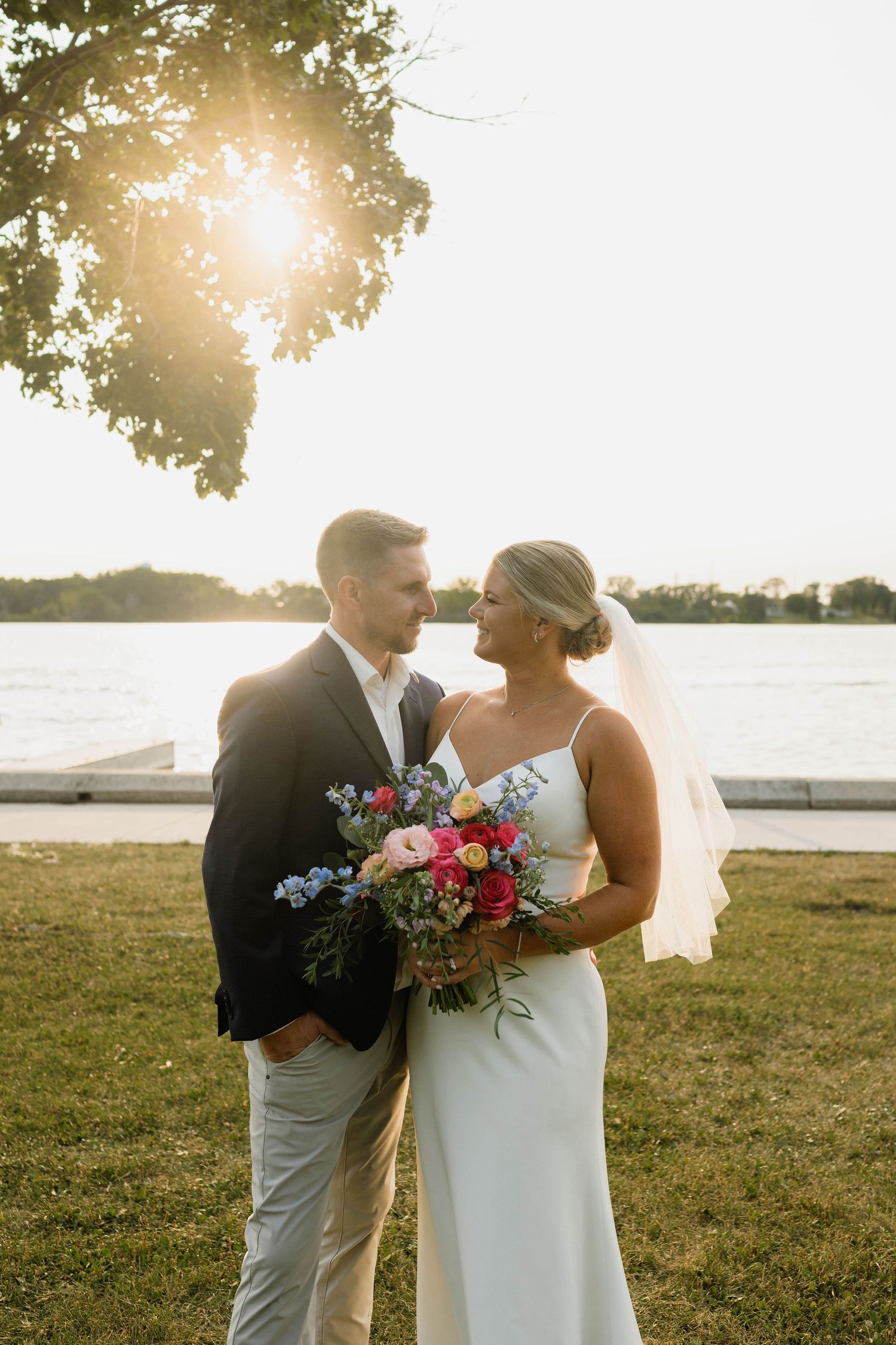 Bride and groom gaze at each other smiling at golden hour near a lake, bride holding a vibrant colorful bouquet.