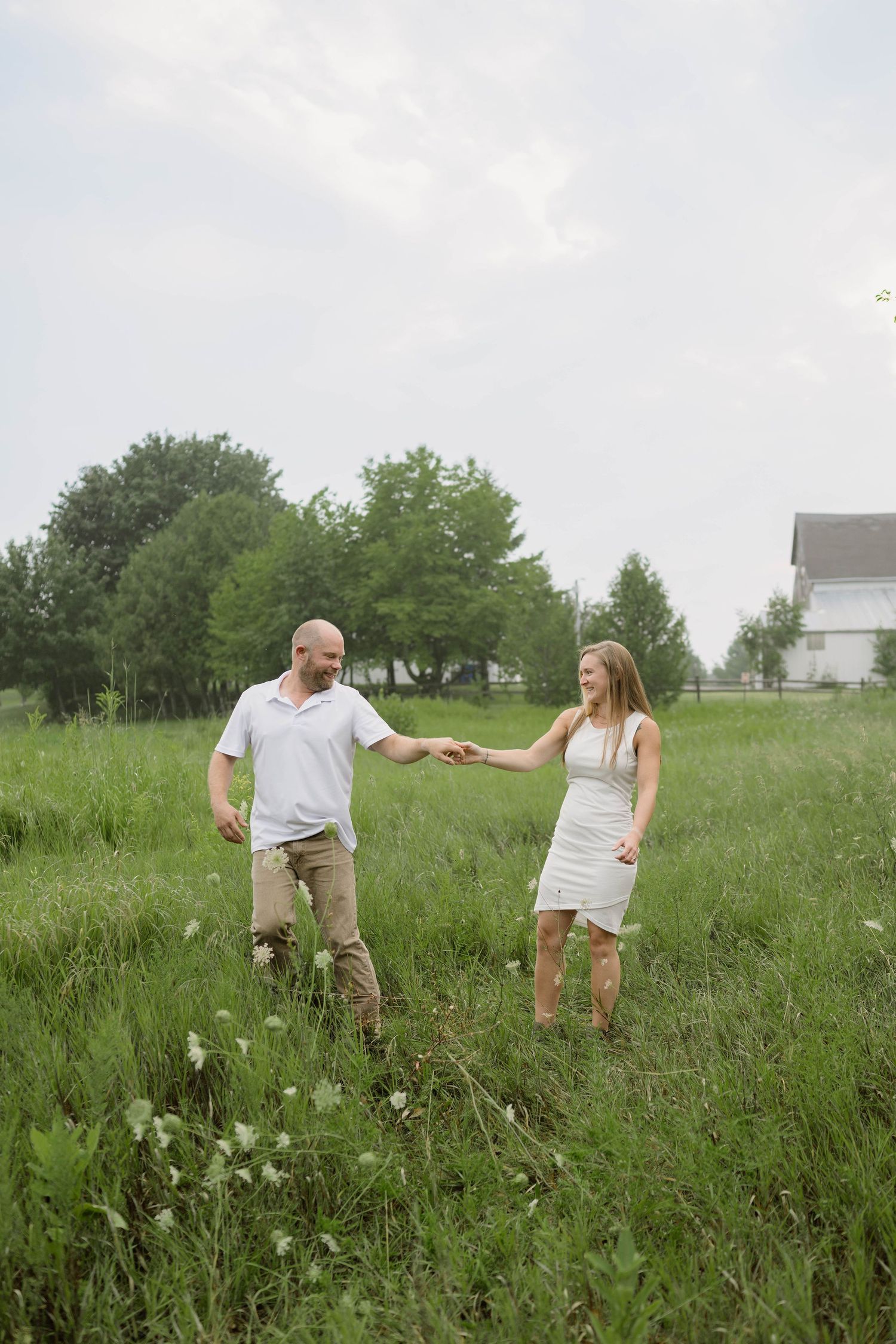 Couple holding hands walking through lush green field, man in white polo and khakis, woman in white dress.