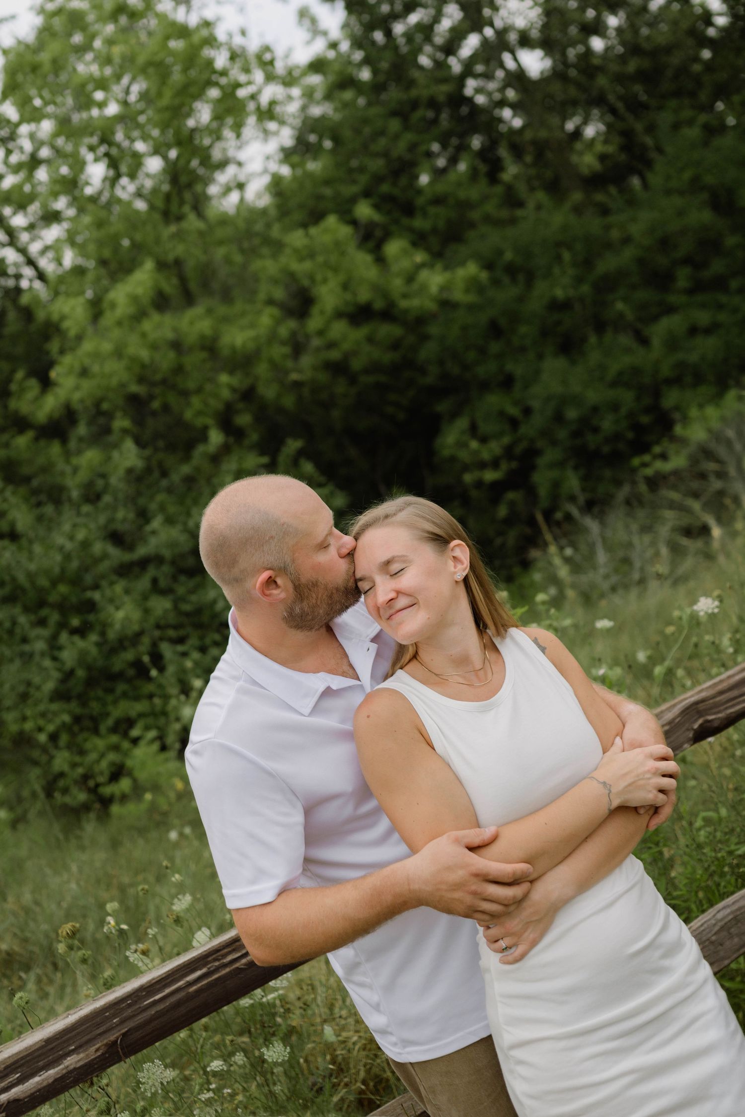 Man hugging and kissing woman on cheek near rustic wooden fence surrounded by lush green foliage.