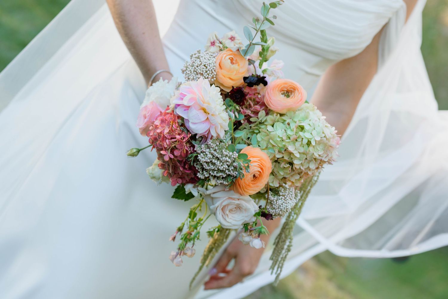 Bride in white wedding dress holding a colorful bridal bouquet with peach ranunculus, pink peonies, and greenery.