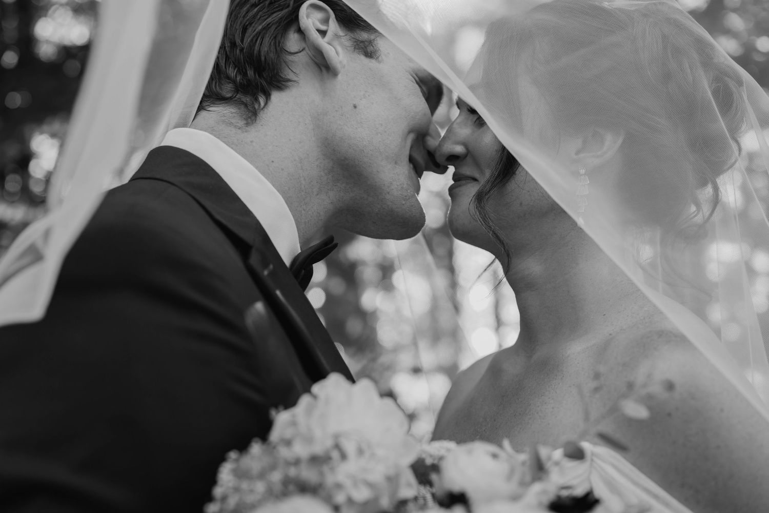 Black and white photo of a bride and groom sharing a tender kiss under a flowing veil on their wedding day.