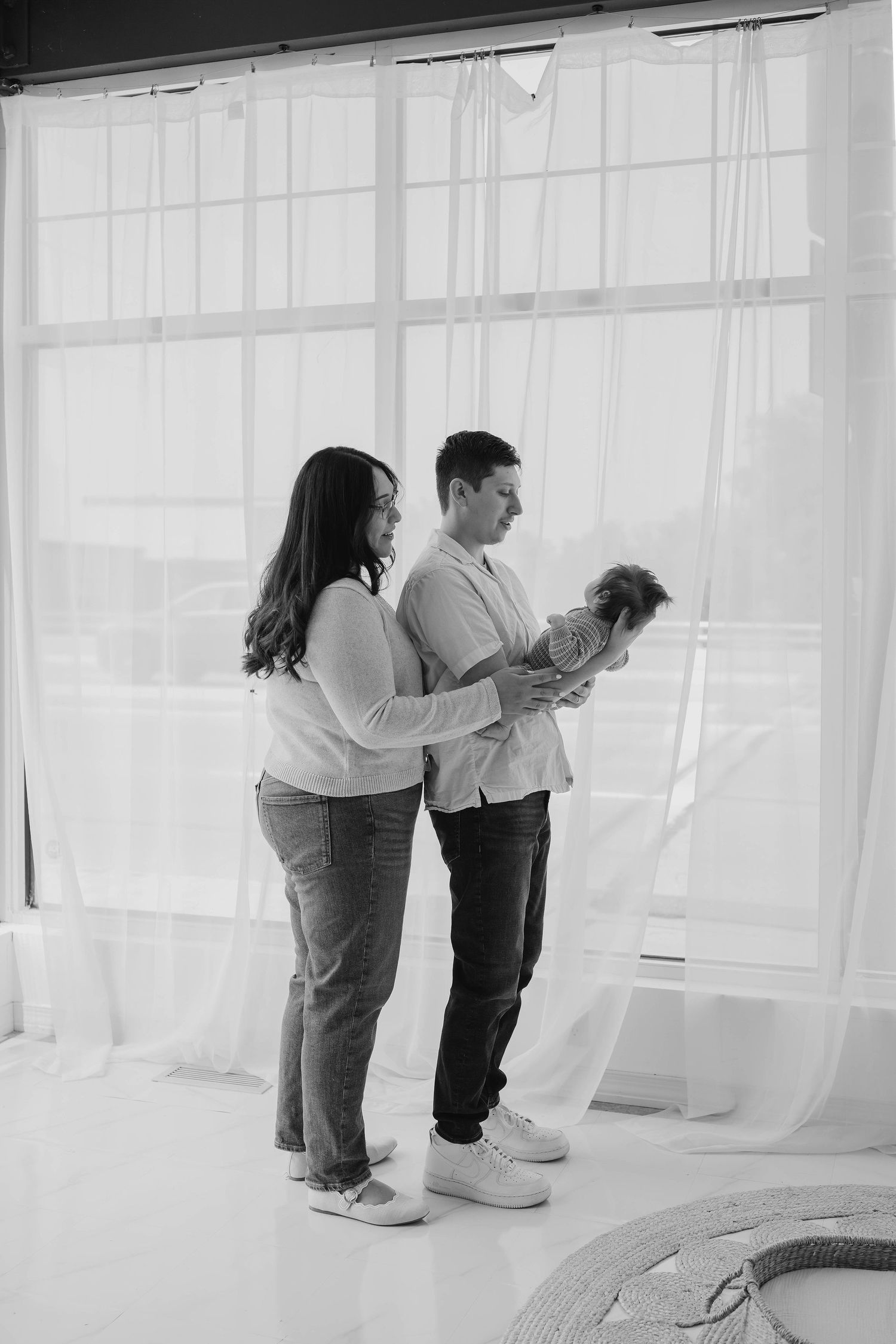 Black and white photo of new parents standing by a bright window, lovingly holding their newborn baby together.