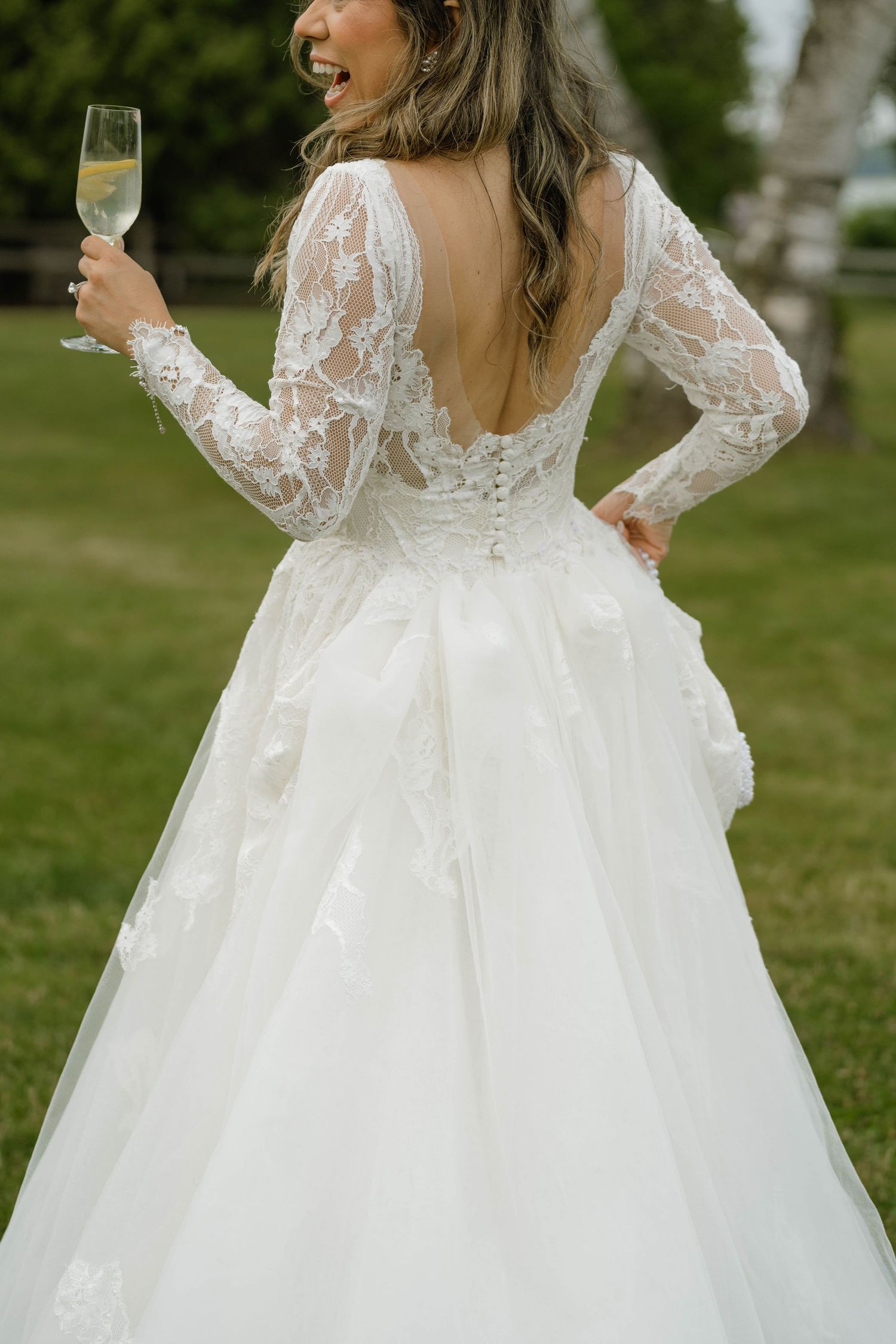 Bride in backless lace long-sleeve wedding gown holding champagne glass on green lawn.