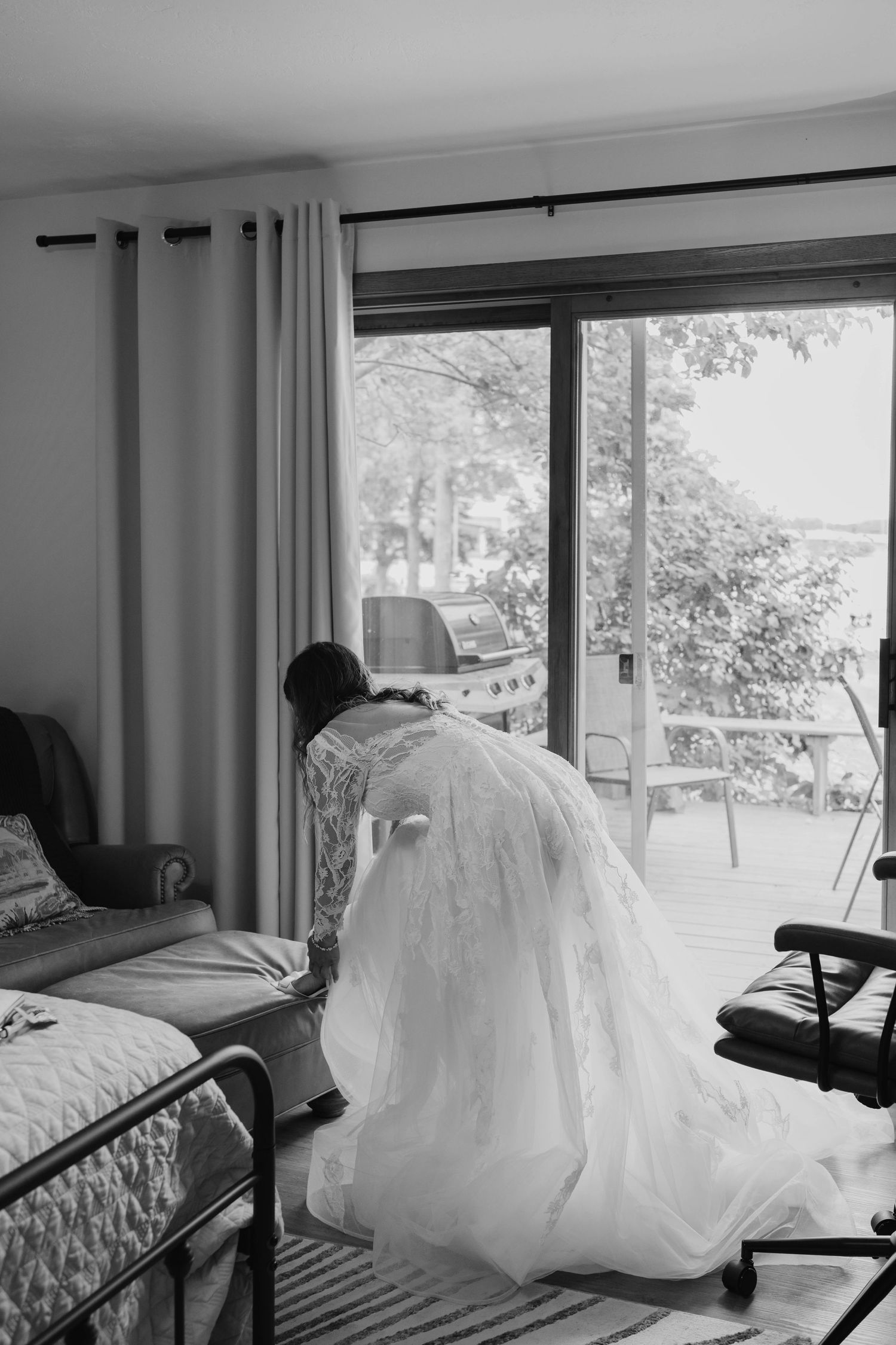 Black and white photo of bride in lace gown sitting by sliding glass door with lake view.