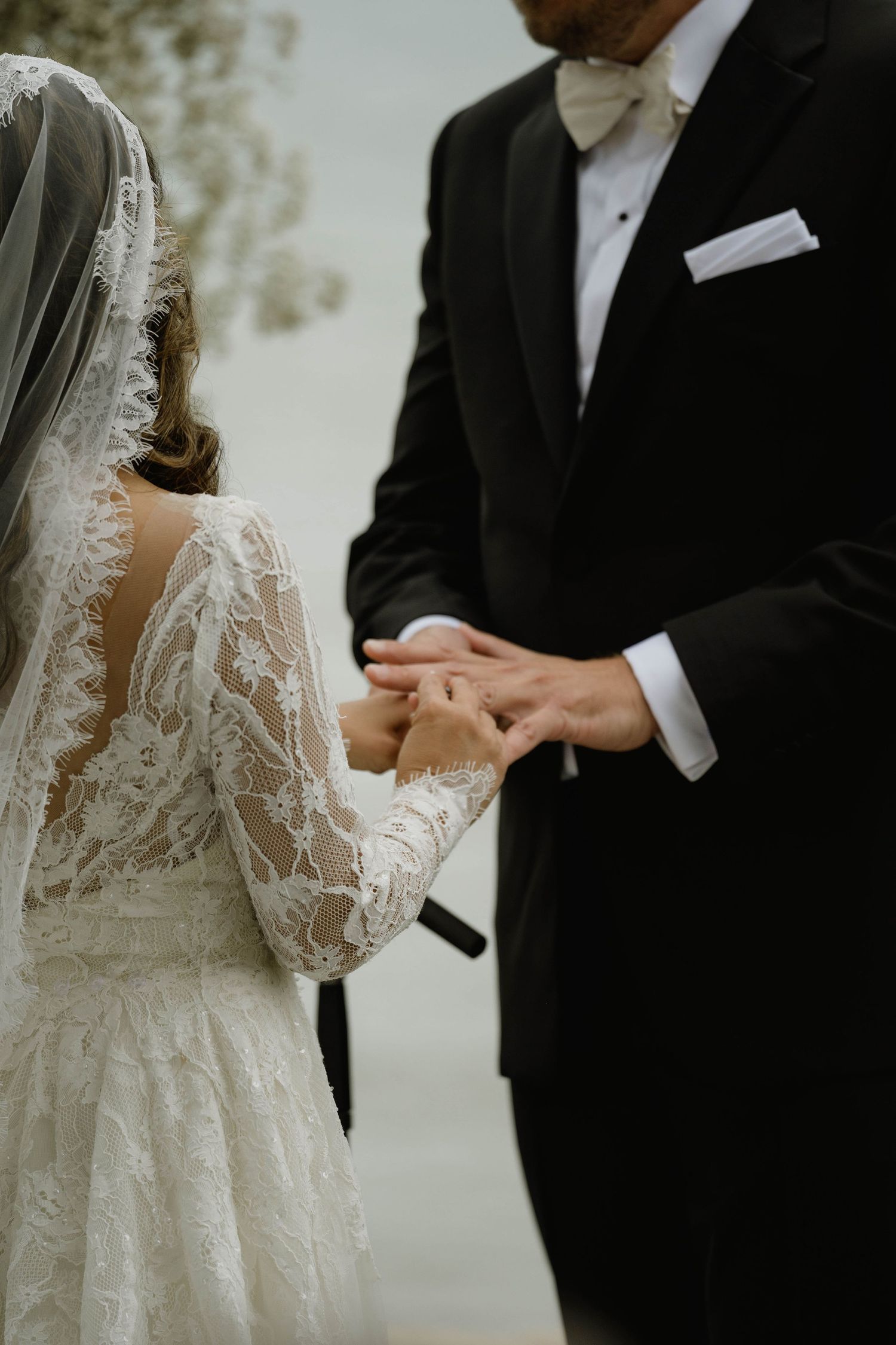 Bride in lace gown places wedding ring on groom's finger during outdoor ceremony.