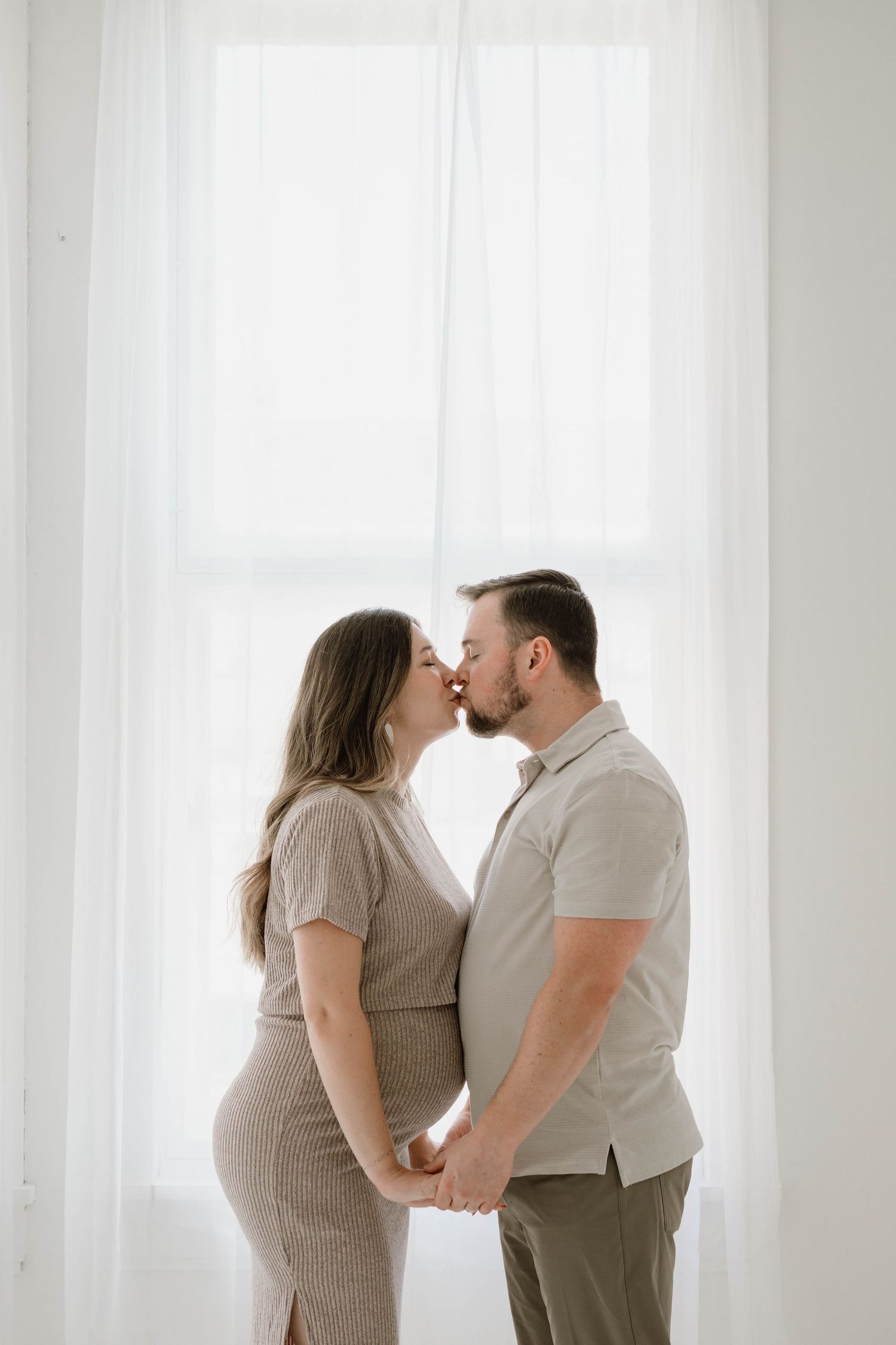 Pregnant couple shares a kiss while holding hands in front of bright white curtained window.