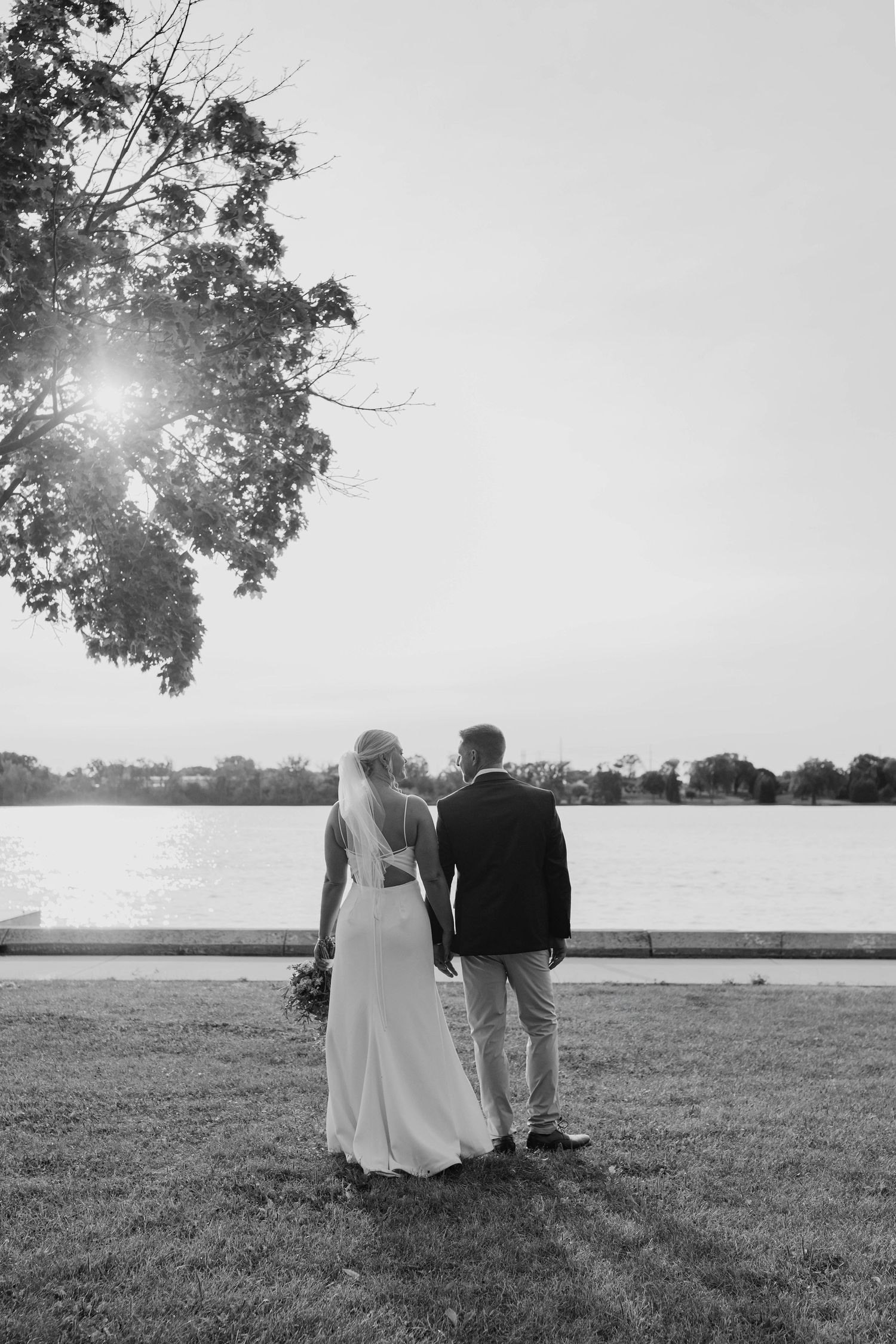 Black and white photo of newlyweds standing under tree gazing at sunlit lake together.