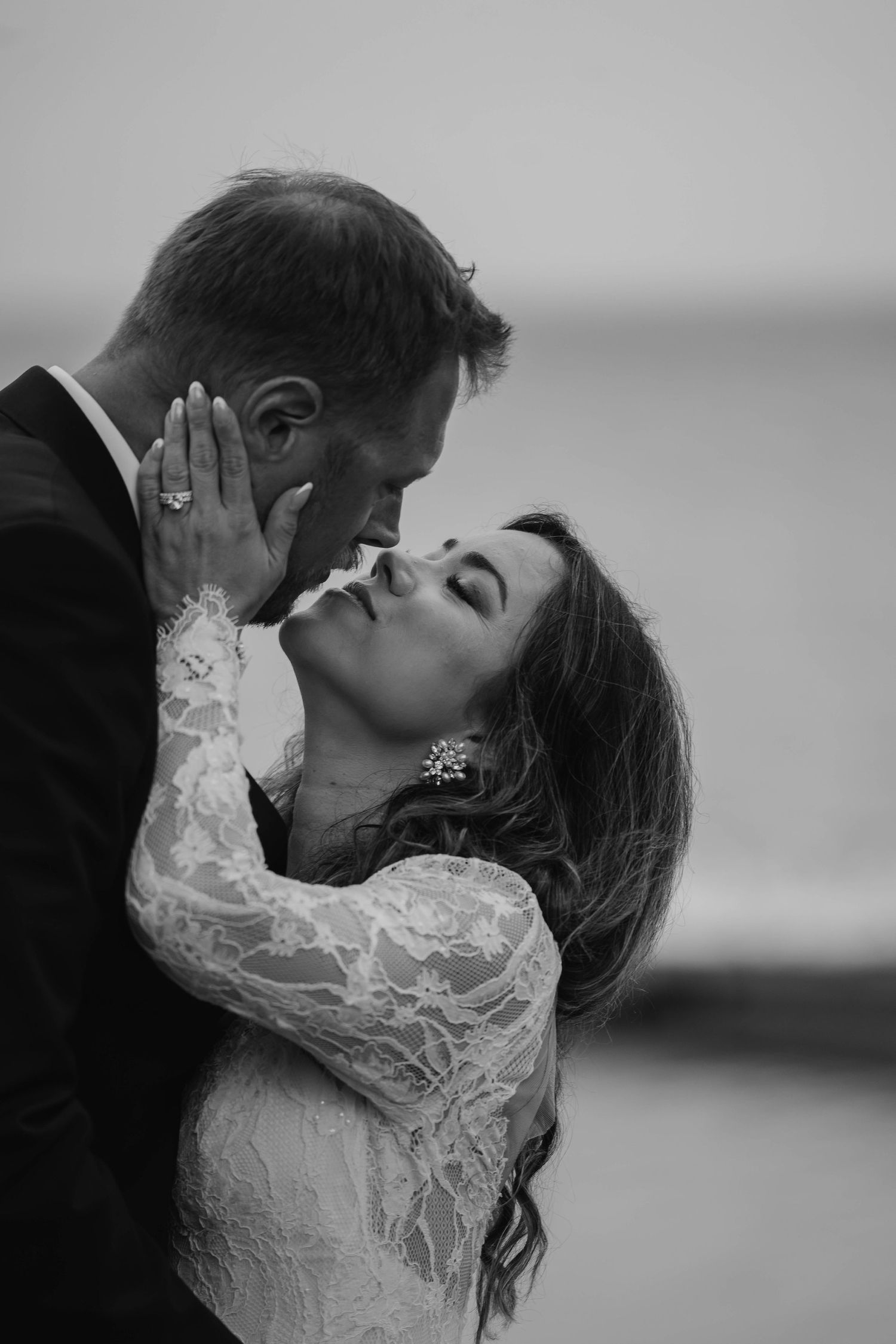 Black and white photo of a bride in a lace wedding dress with her hand on her groom's face as they share an intimate moment by the sea.