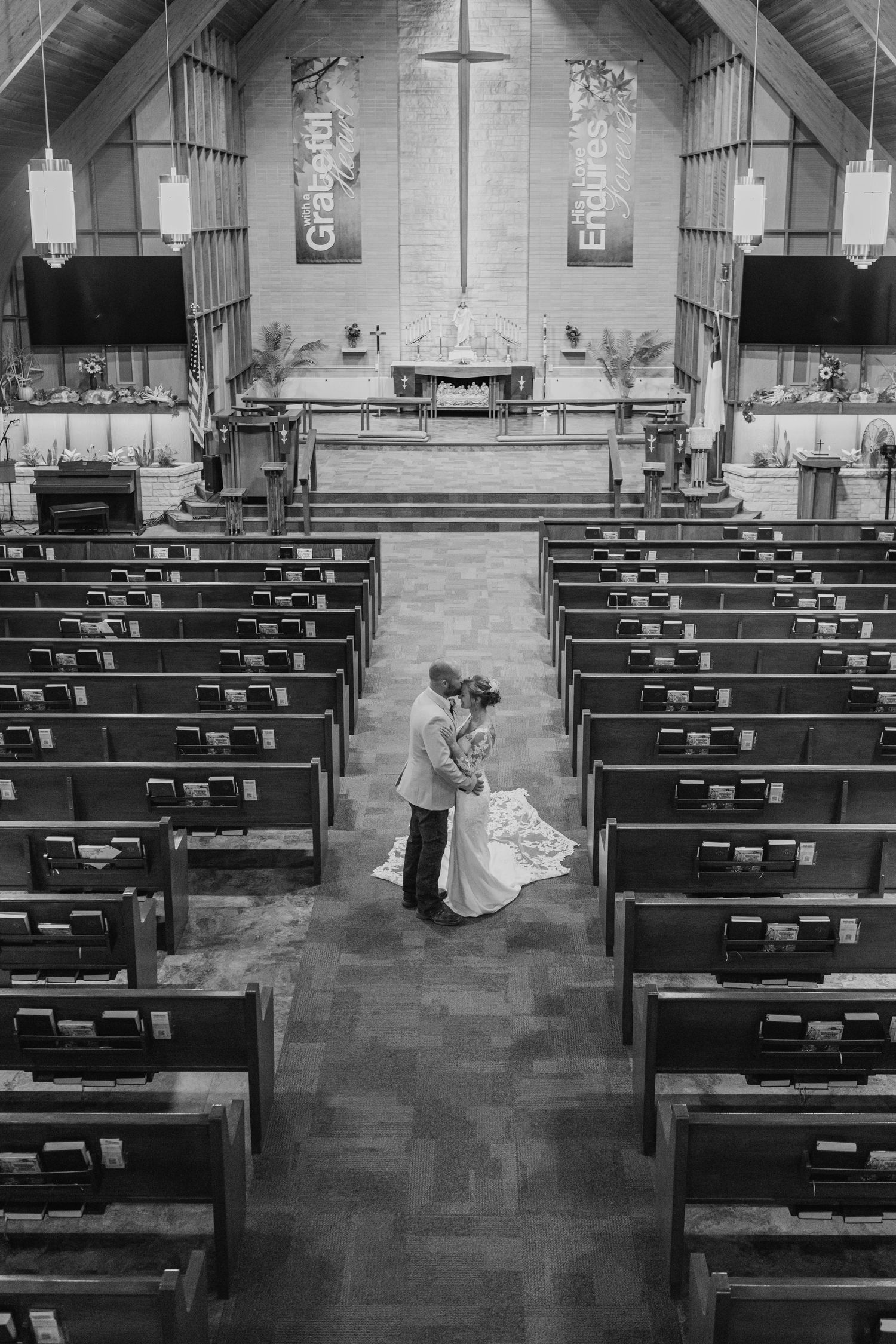 Black and white photo of a bride and groom kissing in the center aisle of an empty church, surrounded by rows of wooden pews.