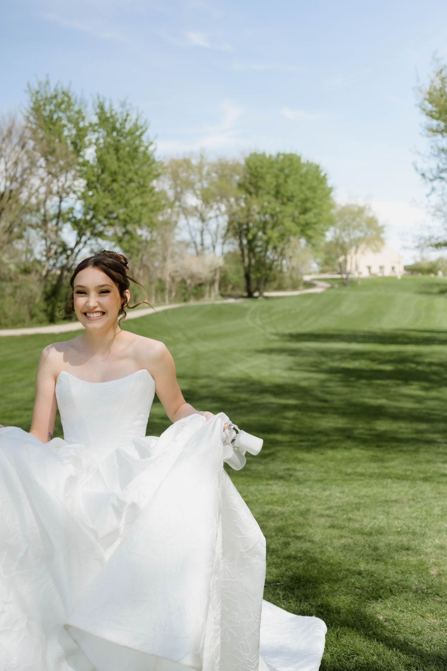 Happy bride in a strapless white wedding gown lifts her skirt while walking across a lush green lawn.