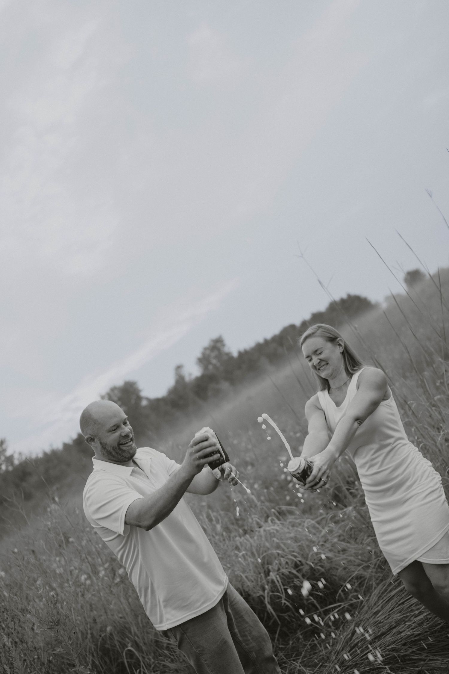 Black and white photo of a couple laughing while opening a champagne bottle in a meadow.